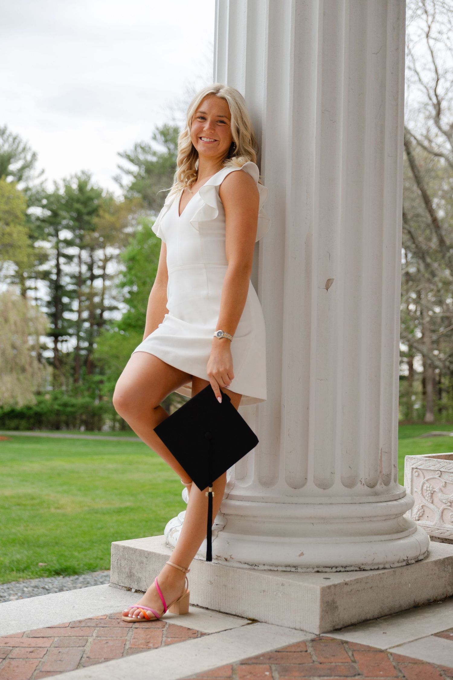 Blonde graduate in white dress leaning against white column holding graduation cap outdoors.