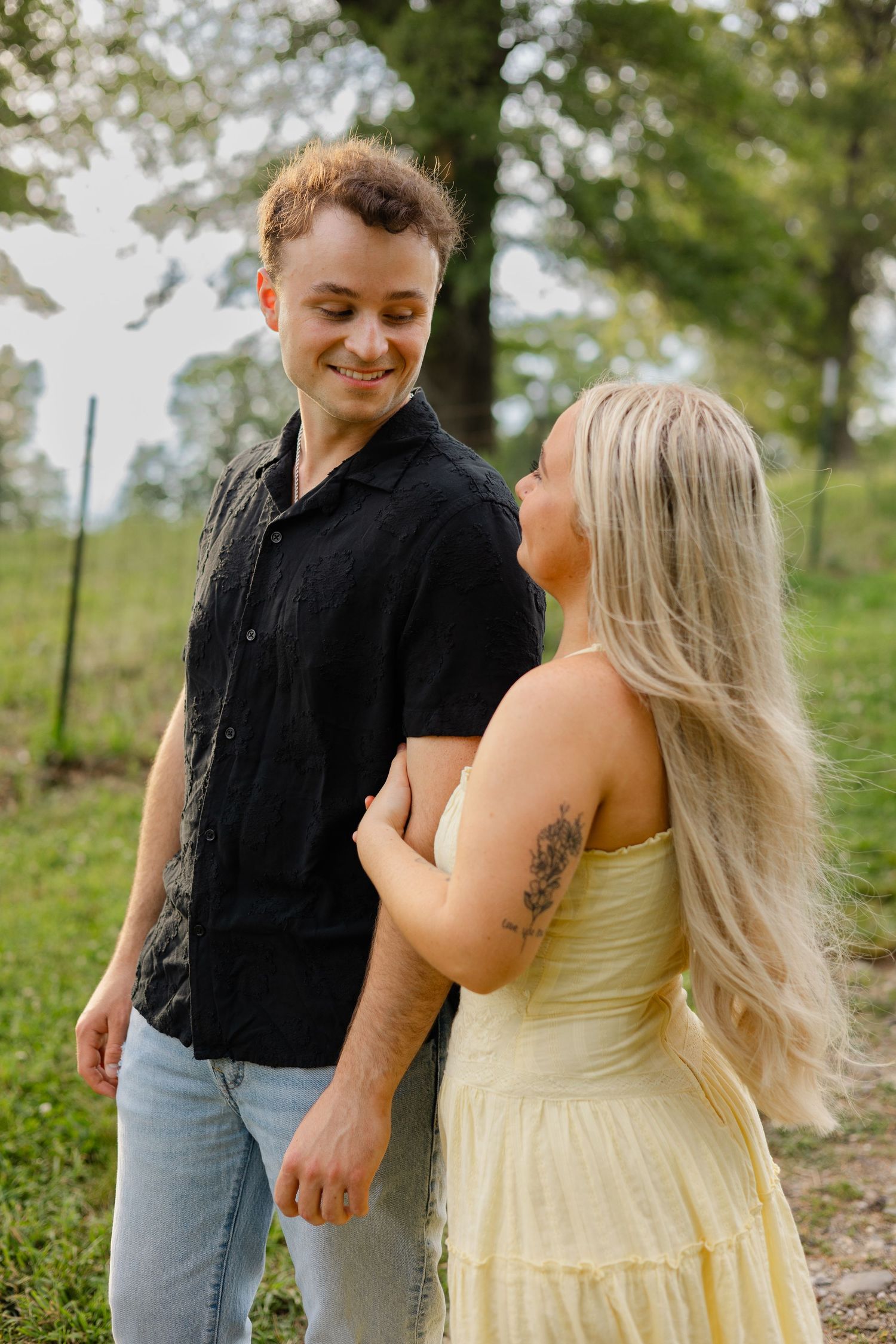 A smiling man in a black shirt and jeans stands with a blonde woman in a yellow dress outdoors.