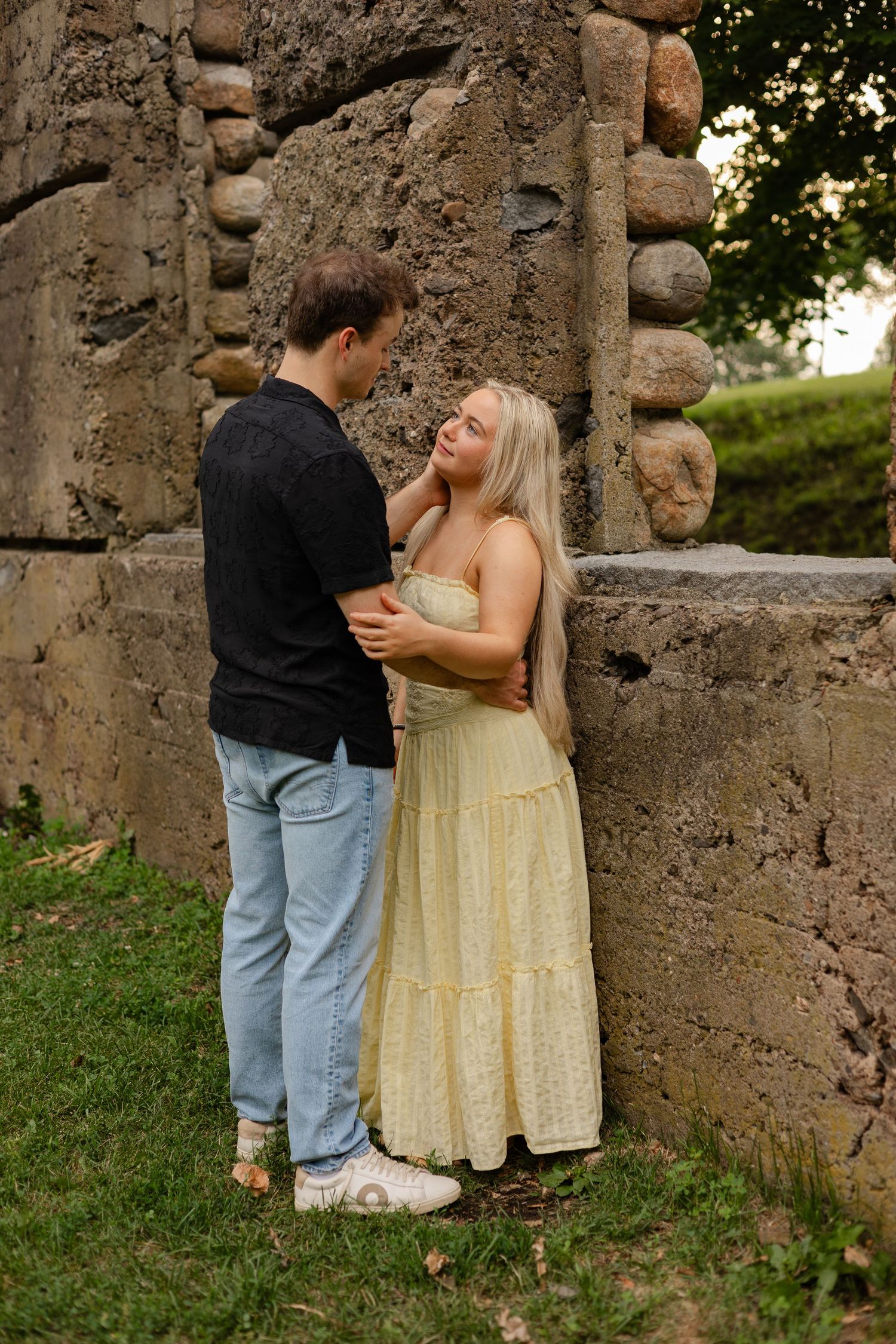 A couple in casual attire sharing a tender moment against a rustic stone wall in a park.