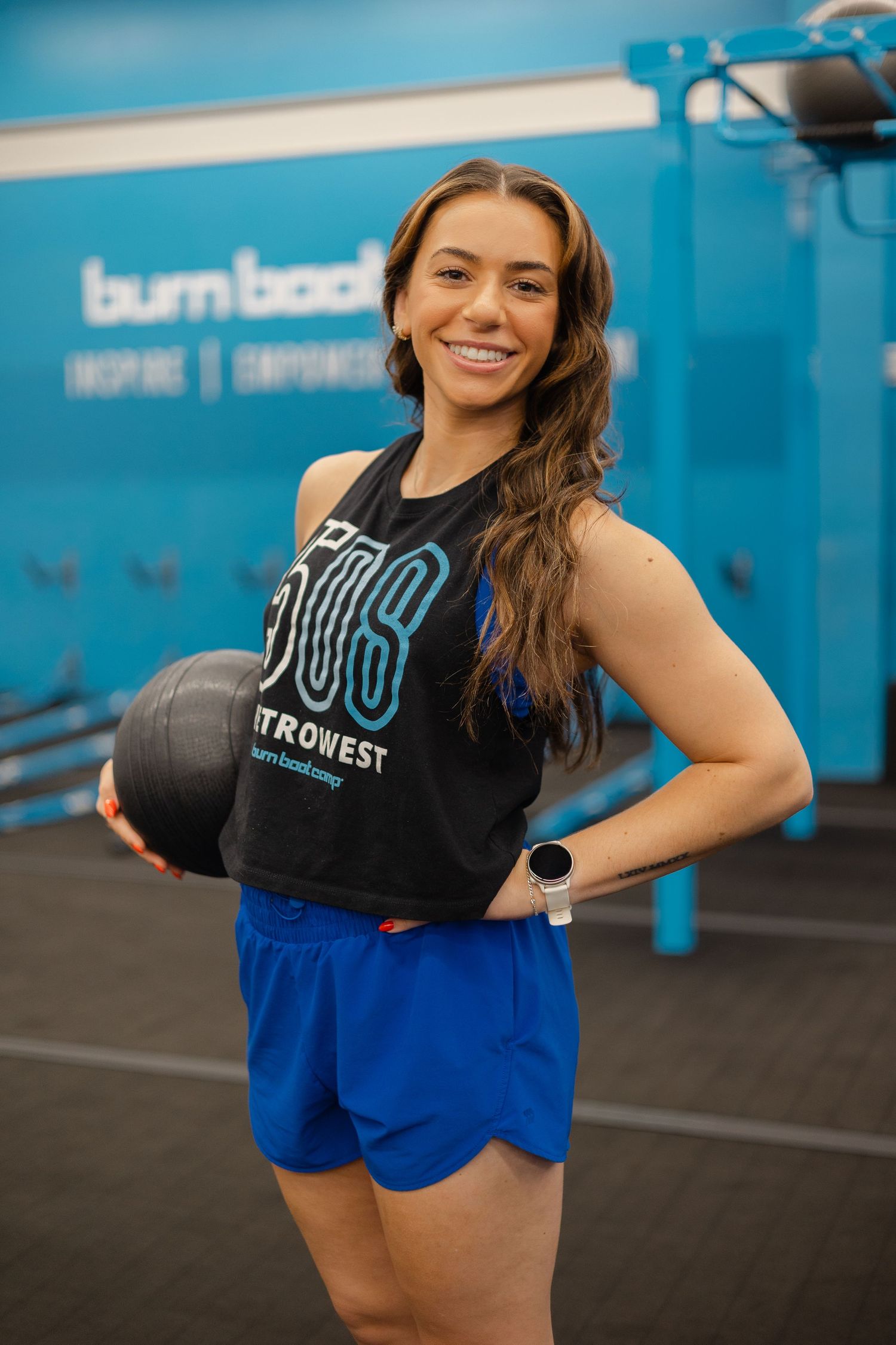 A smiling woman in a black tank top and blue shorts holds a medicine ball at a Burn Boot Camp gym.