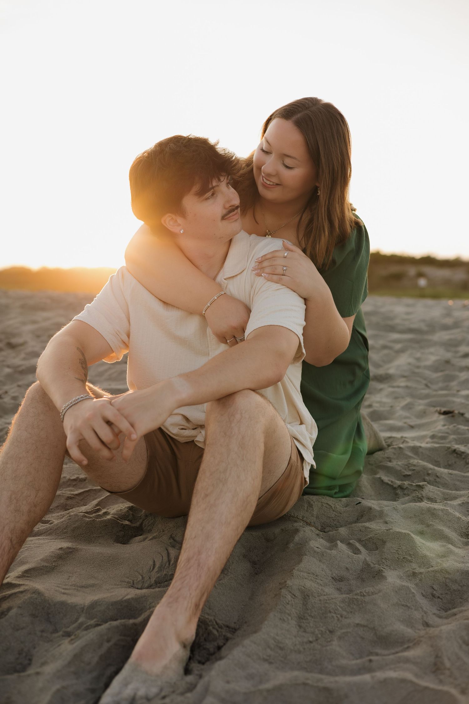 Couple sitting on sandy beach at golden sunset, woman in green dress embracing man from behind.