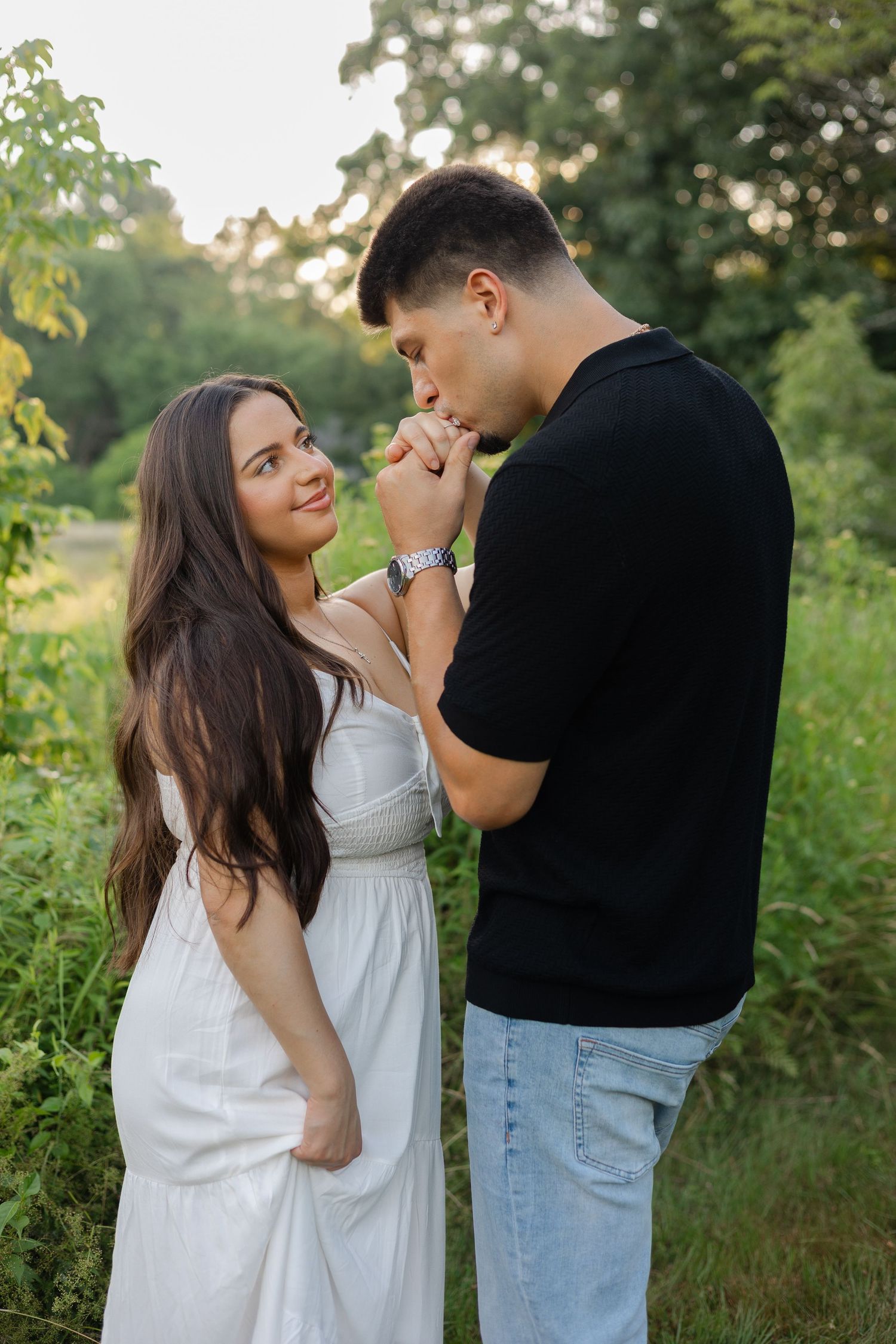 A man in a black shirt kisses a woman's hand outdoors, as she smiles in a white dress amid lush greenery.