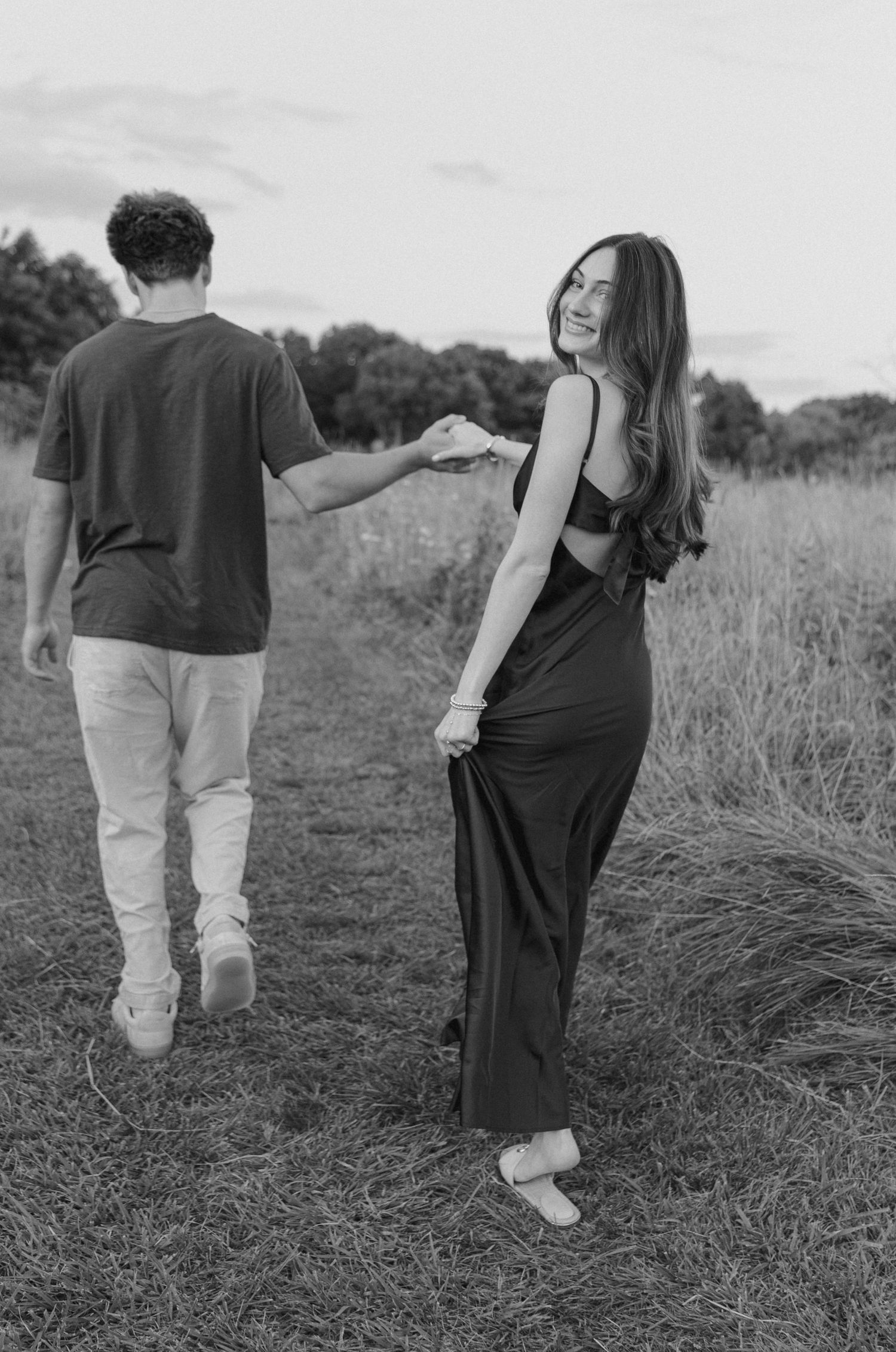 Black and white photo of a couple holding hands walking through a field, woman in maxi dress smiling back.