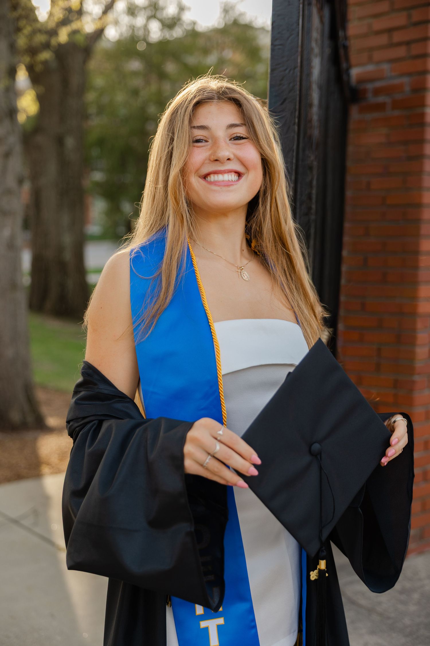 Smiling young woman in graduation cap, black gown, and blue sash poses outdoors near brick building.