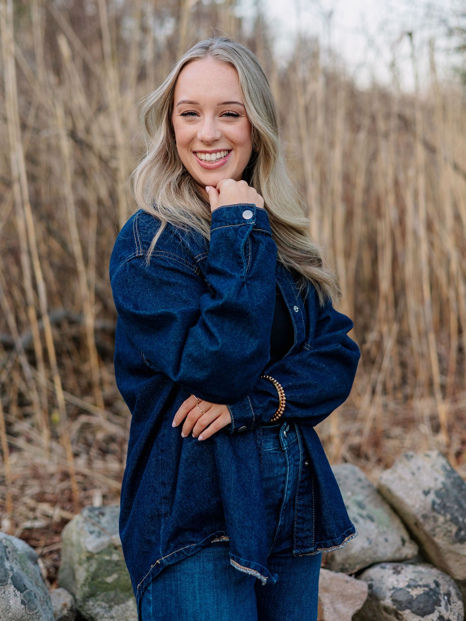 Smiling blonde woman in dark denim jacket and jeans posing outdoors among rocks and tall grass.