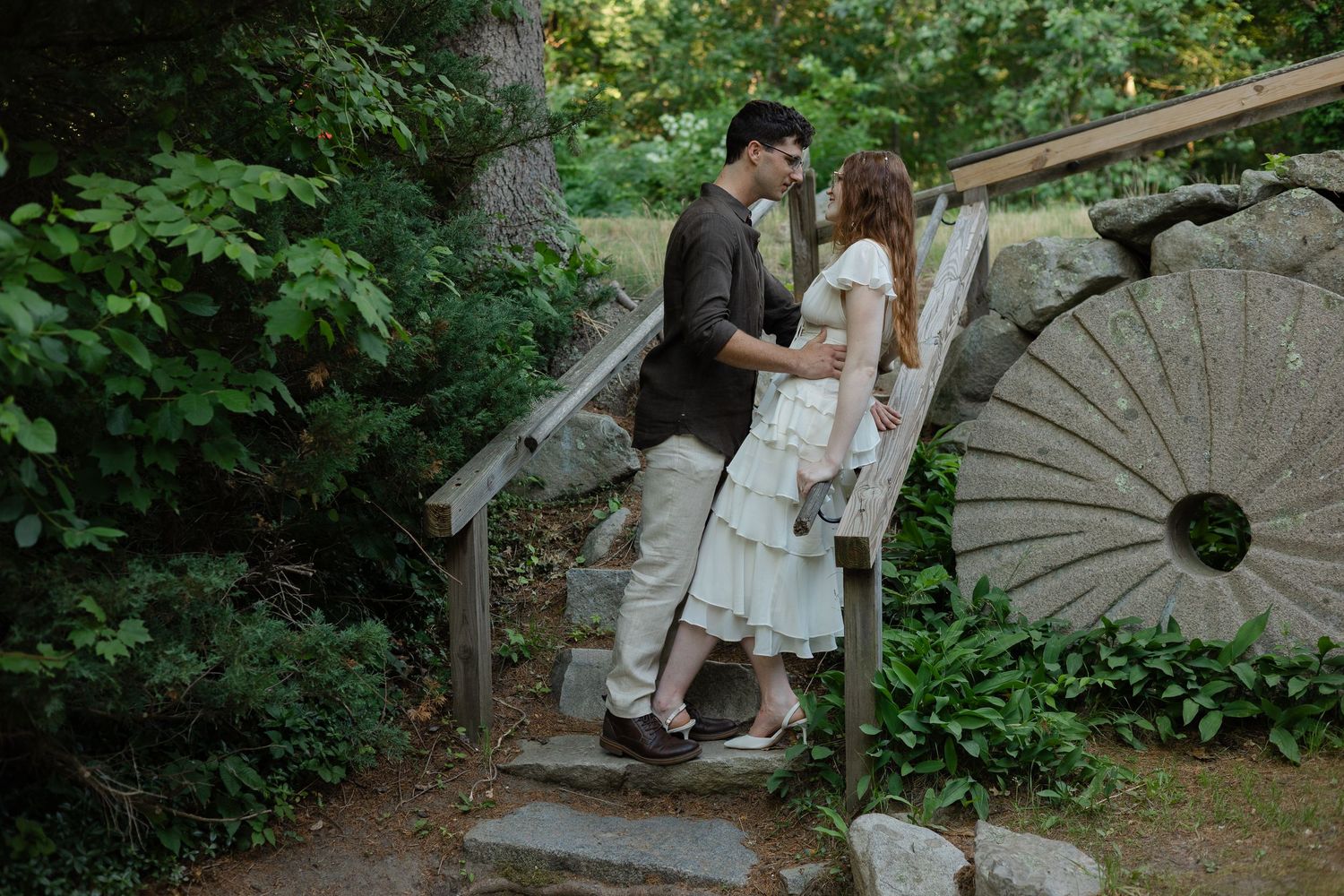 Couple sharing a romantic moment on rustic stone steps surrounded by lush greenery near a decorative millstone.