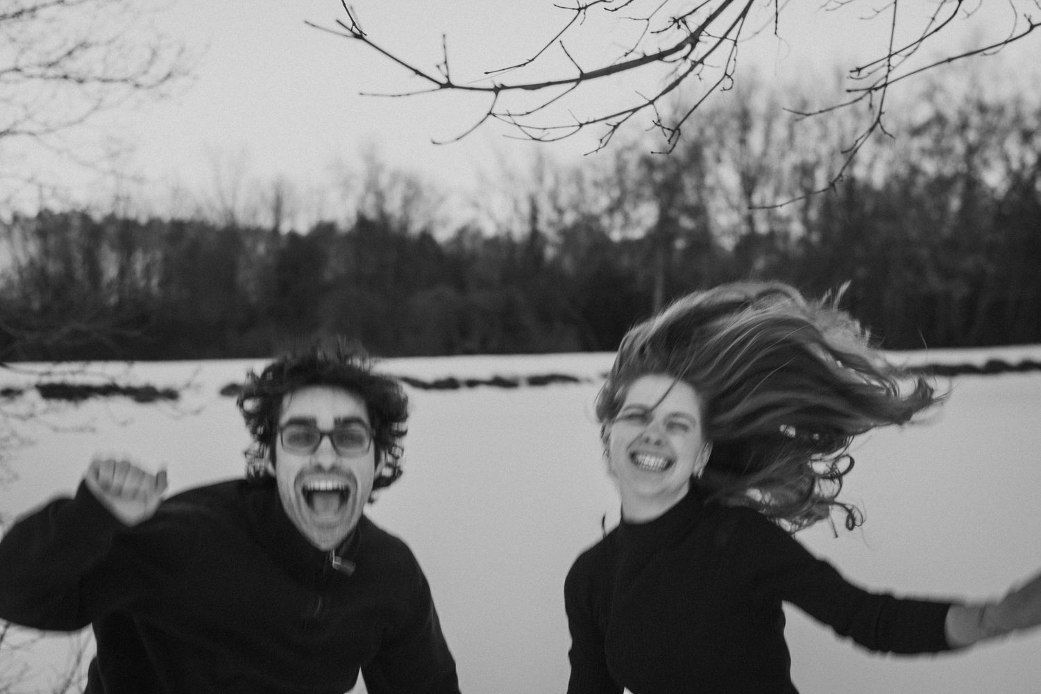 Black and white photo of laughing couple jumping joyfully outdoors near frozen winter landscape.