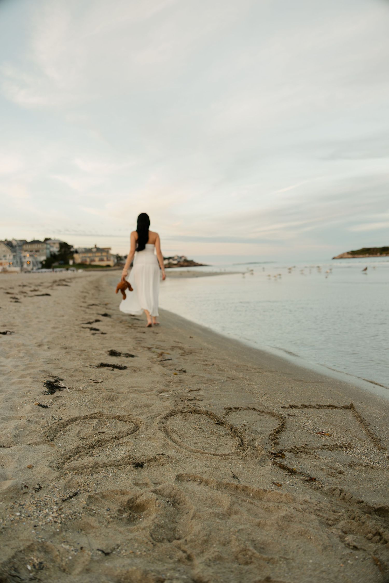 Woman in white dress walking barefoot along sandy beach at dusk, with writing in sand and calm ocean waters.