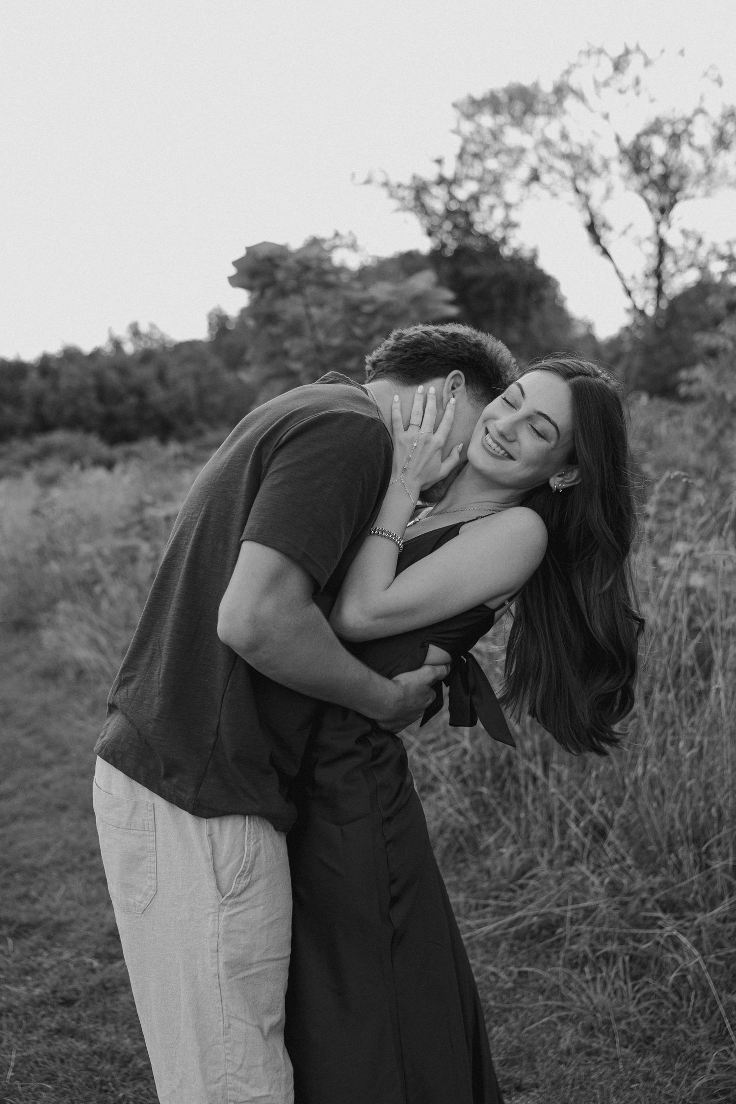 Black and white photo of a couple embracing romantically in a field, woman smiling joyfully as man nuzzles her neck.