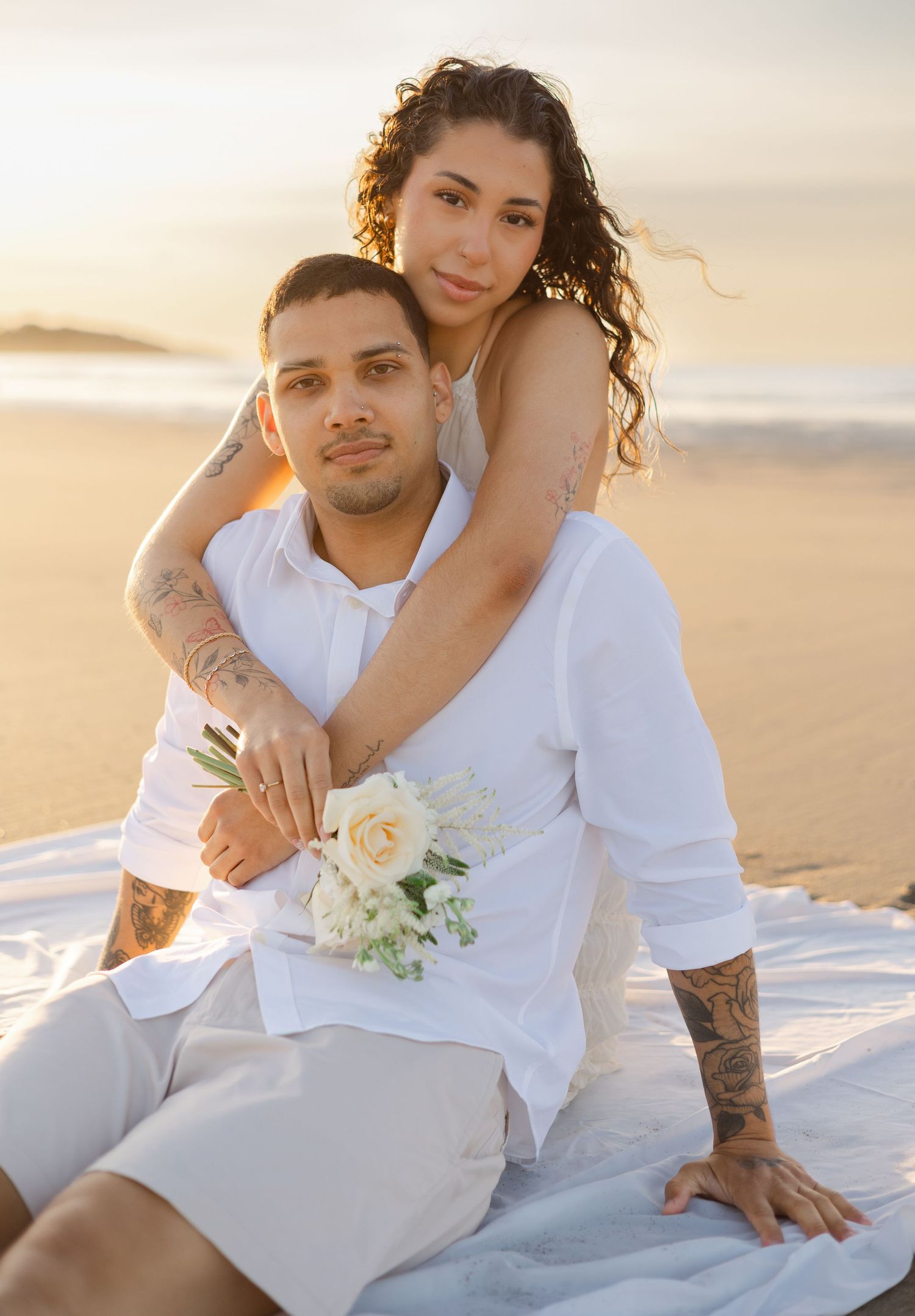 Couple in white attire poses on beach at golden hour, woman holding white roses while embracing man from behind.