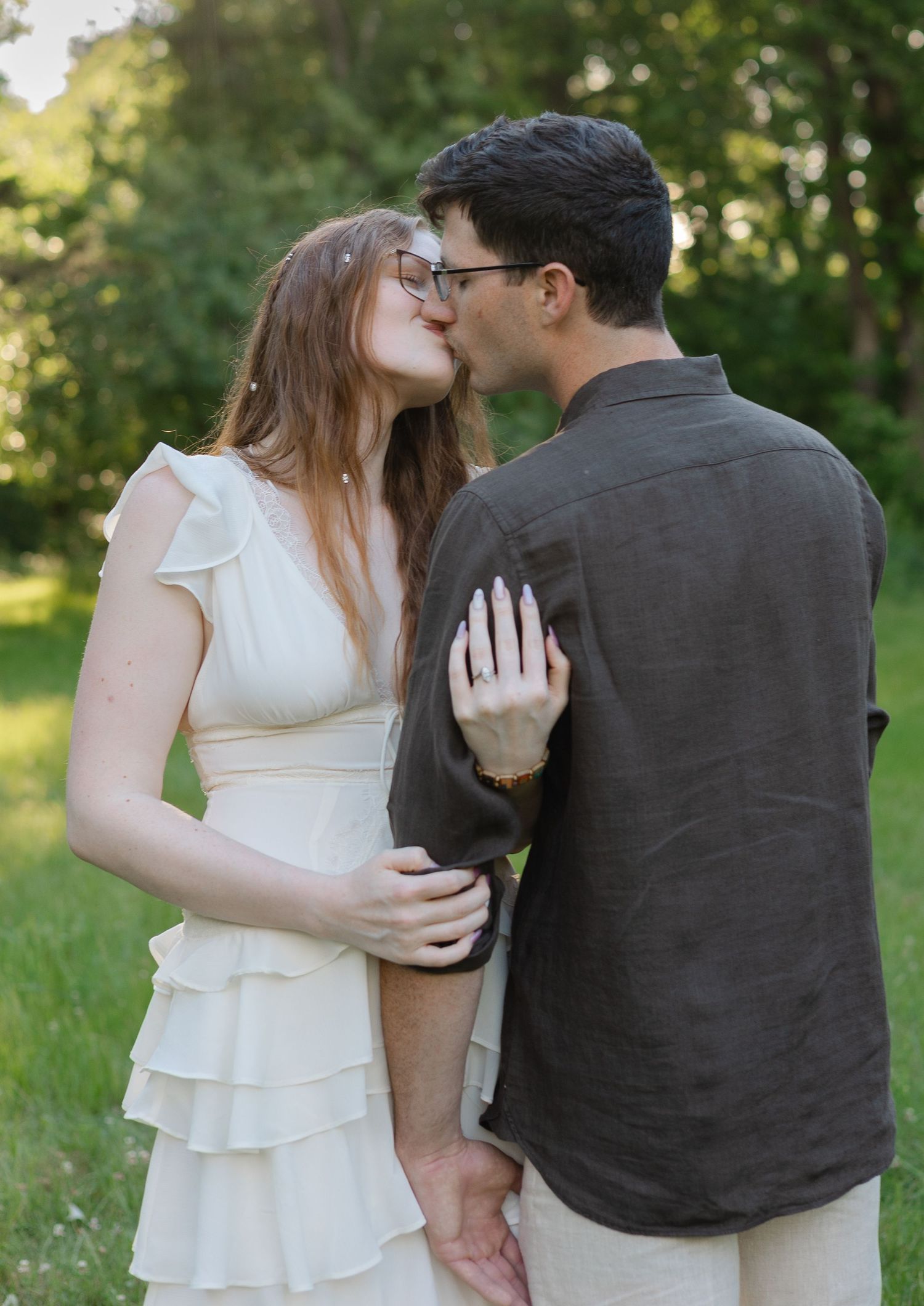 Couple sharing a romantic kiss outdoors, woman in white ruffled dress showing engagement ring, green trees background.