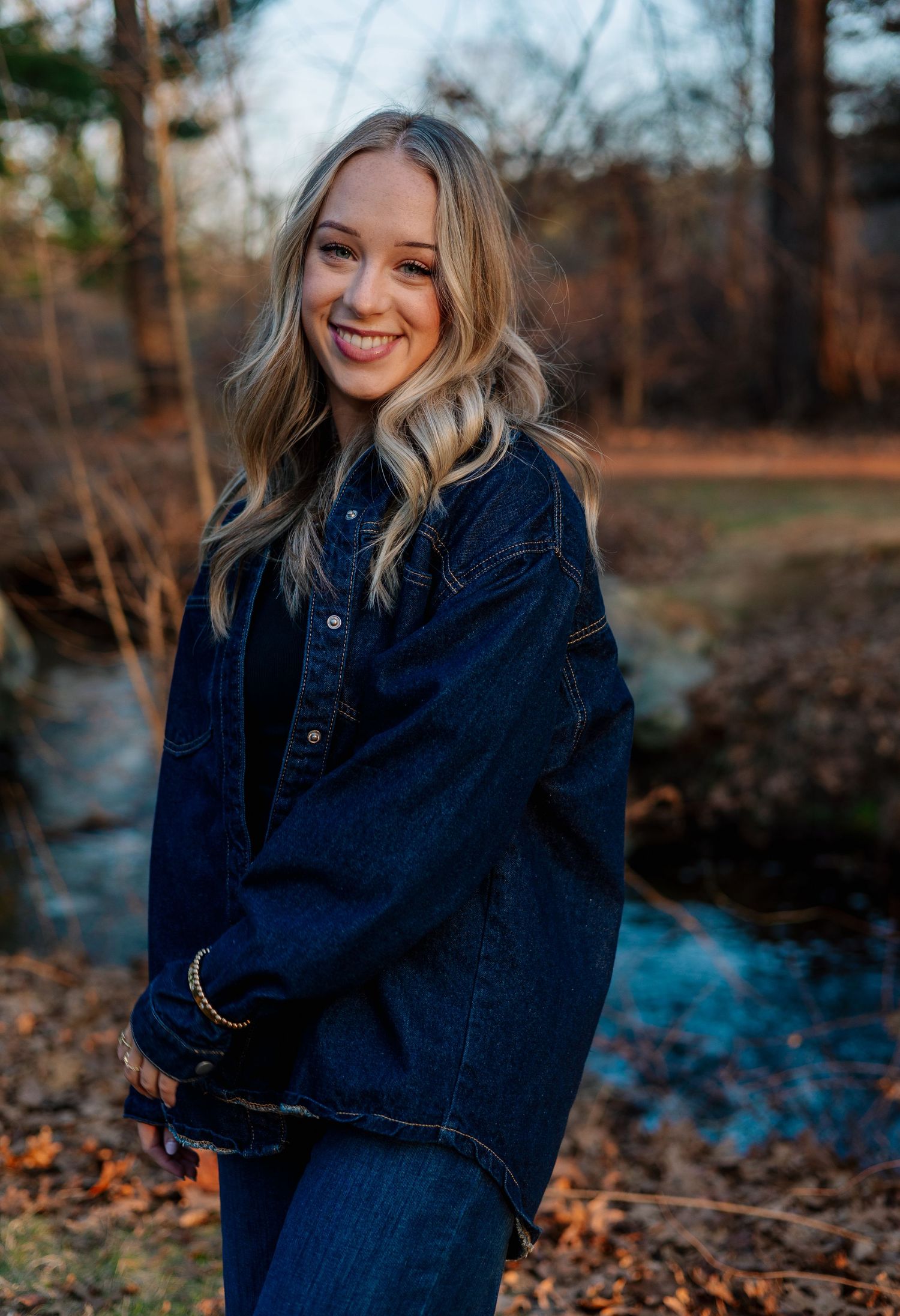 Smiling blonde woman in oversized dark denim jacket posing outdoors near a creek in autumn.