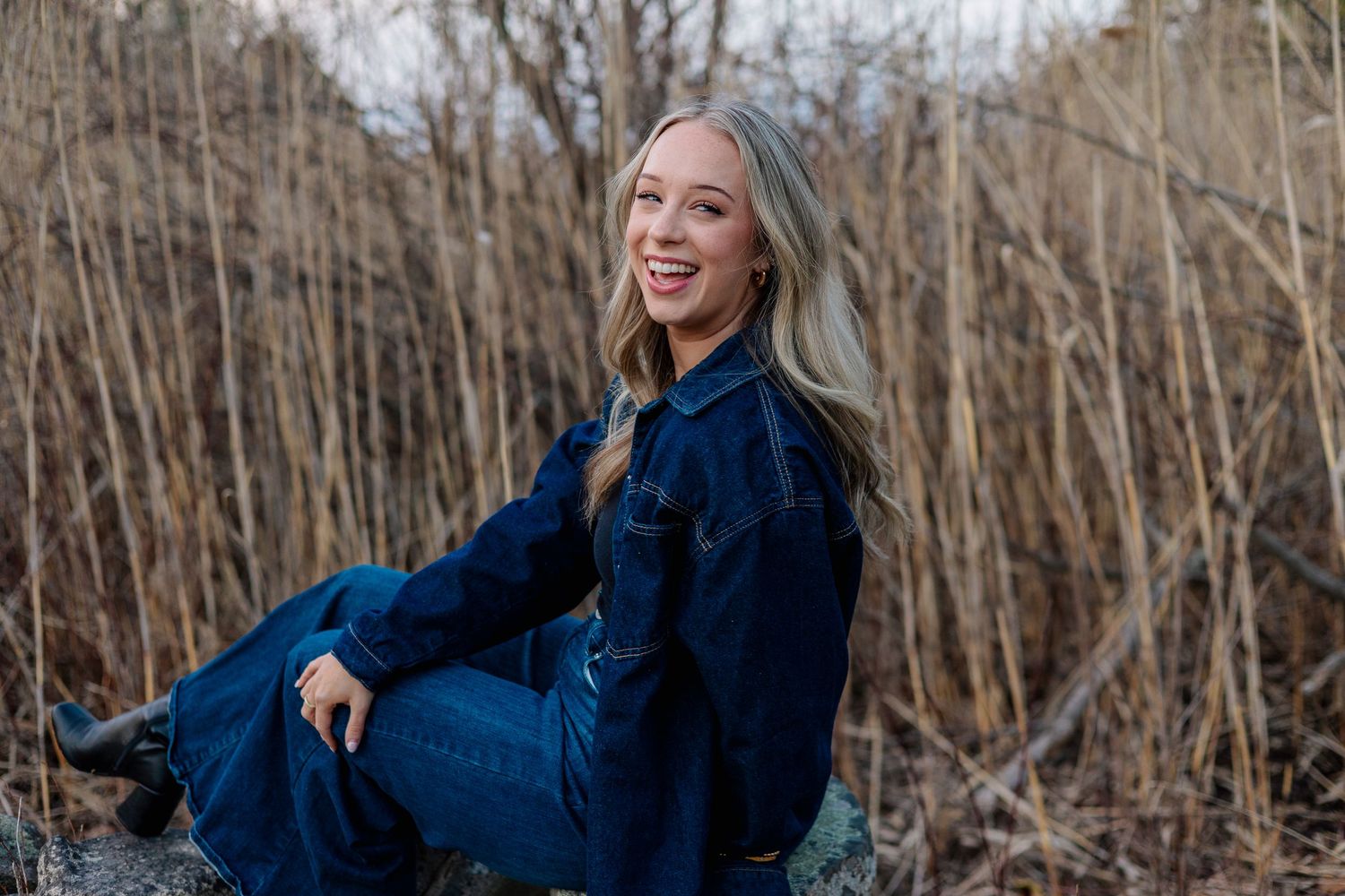 Laughing blonde woman in dark denim outfit sitting on rocks surrounded by tall dried brush.