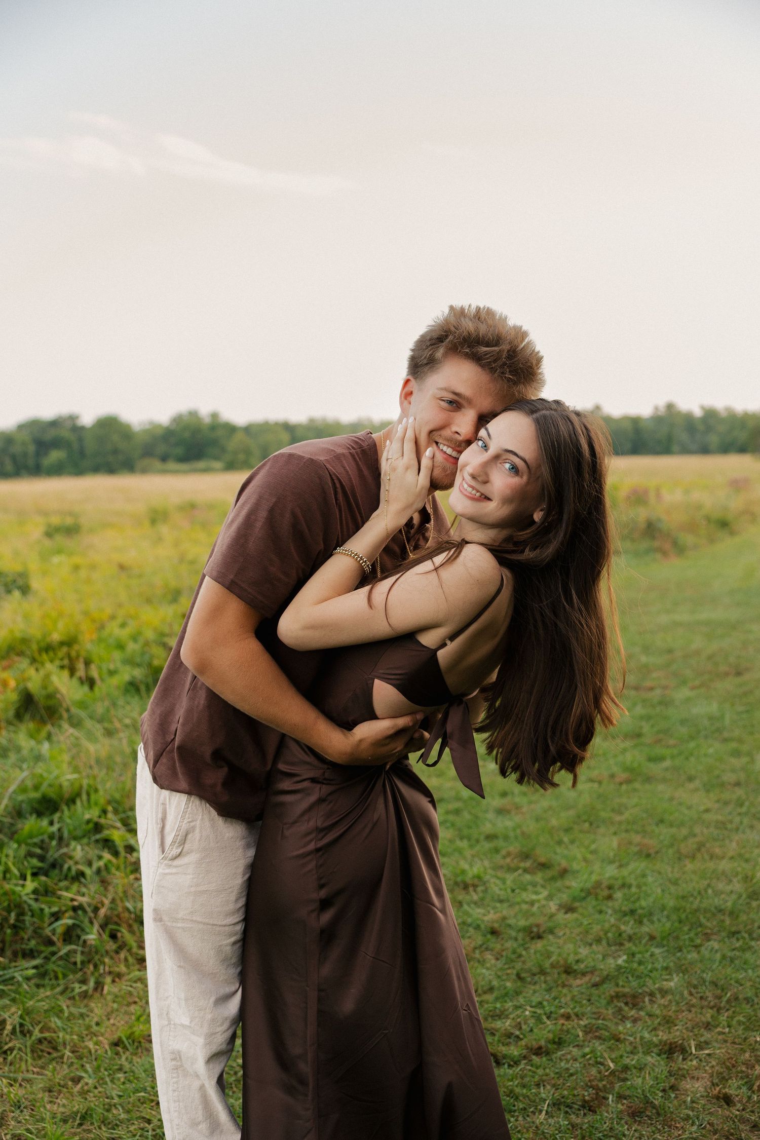A young couple embracing in a green field, wearing brown outfits, smiling warmly in a romantic outdoor setting.