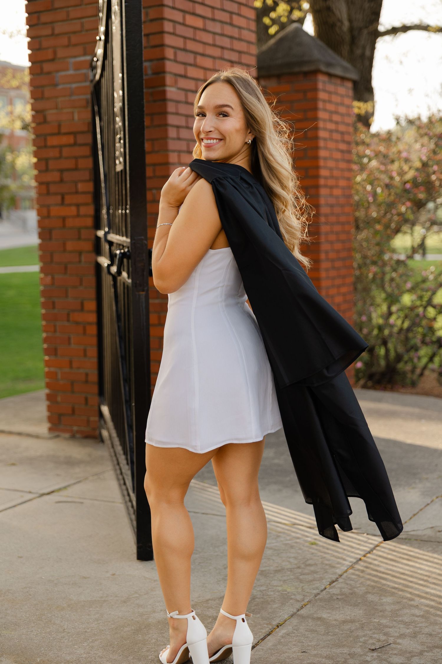 Smiling blonde woman in white mini dress and heels, holding black coat, posing by brick gate outdoors.