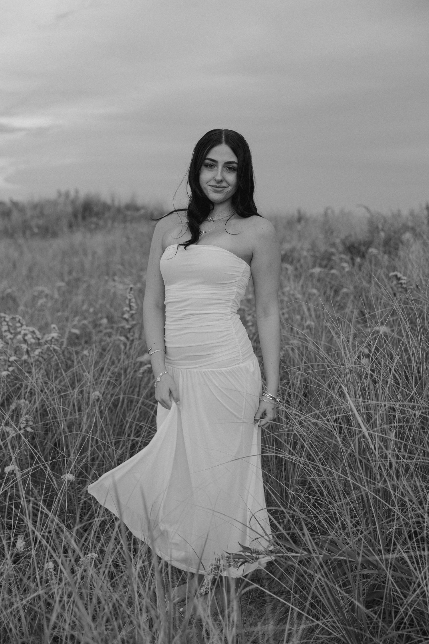 Black and white photo of a young woman in a white strapless dress standing in a field of tall grass.