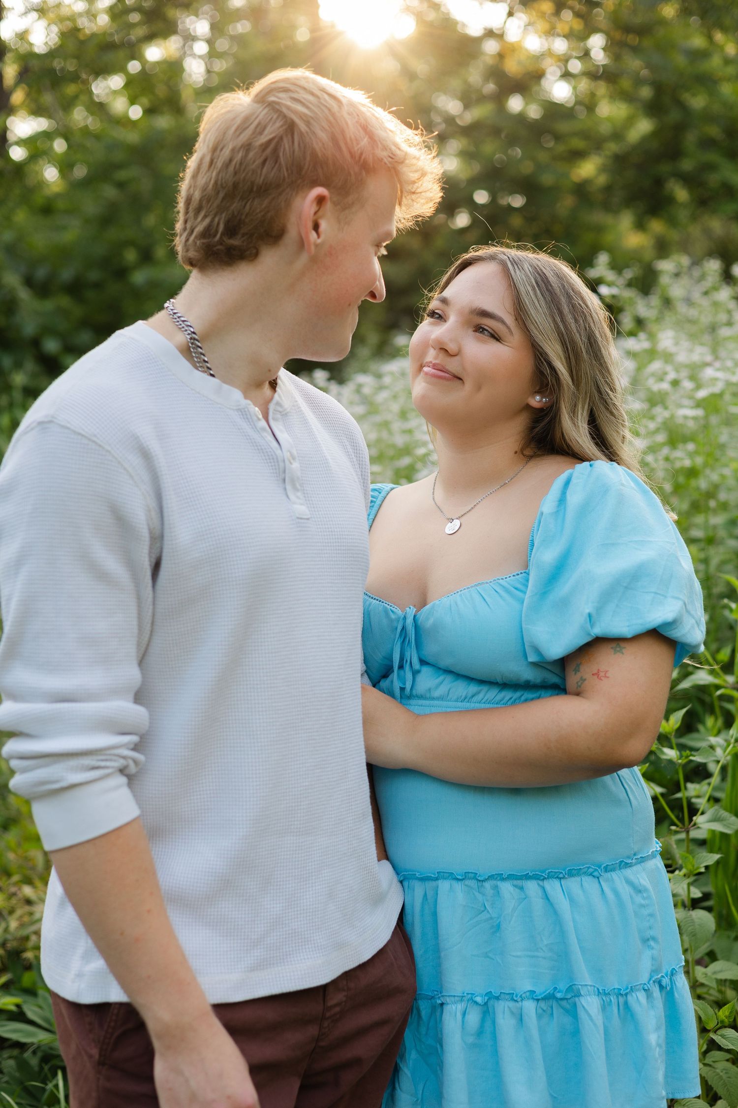 A young couple gazes at each other lovingly outdoors, she in a blue dress, he in a gray shirt, with sunlit greenery behind.
