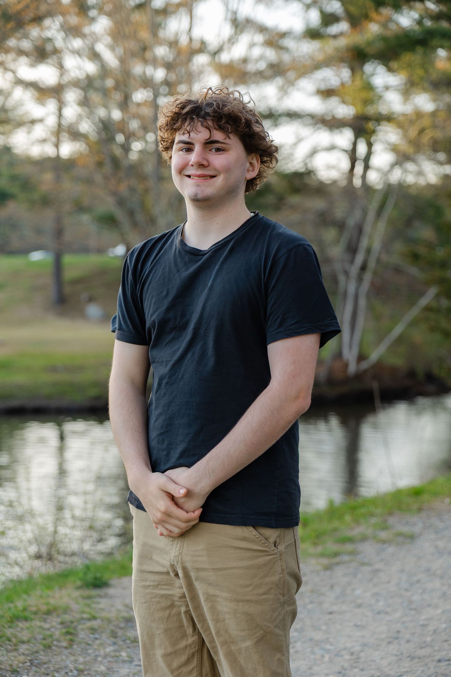 Young man with curly hair in black t-shirt and khaki pants smiling outdoors near a pond with trees.