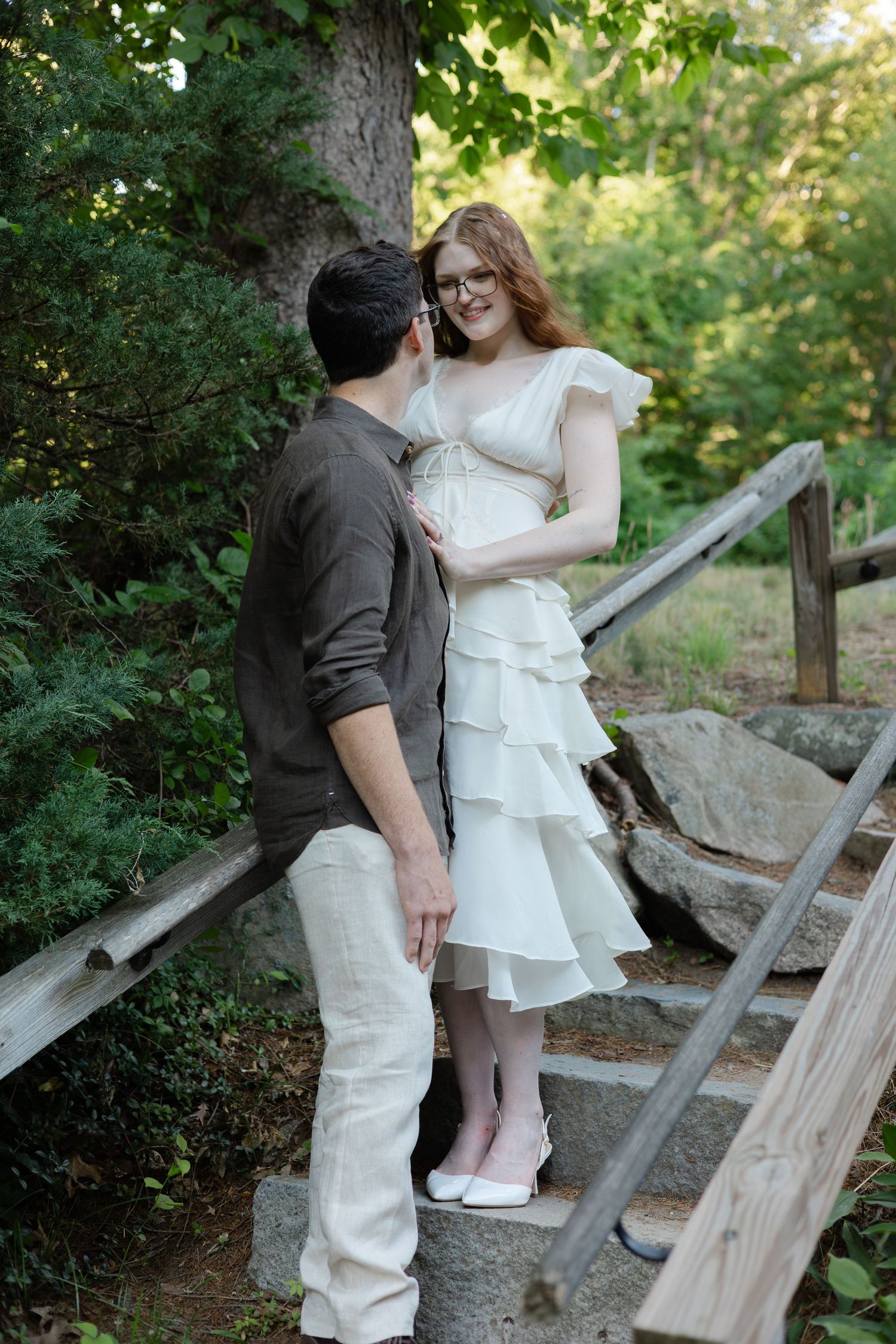 Couple in romantic outdoor setting, woman in white tiered dress on stone steps, man in gray shirt gazing at her.