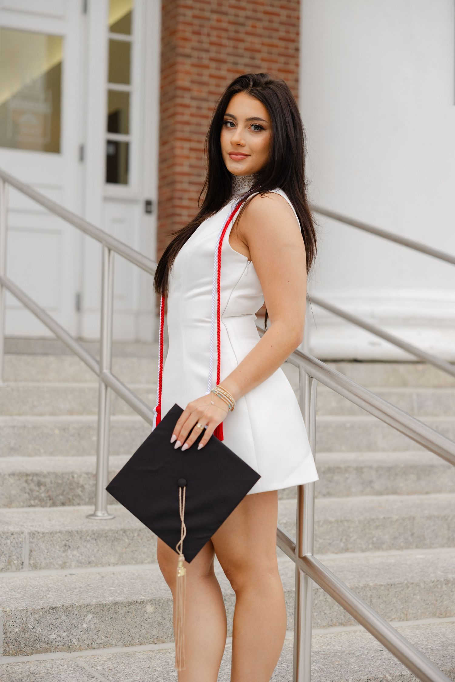 Smiling female graduate in white dress holding graduation cap on university steps with red tassels.
