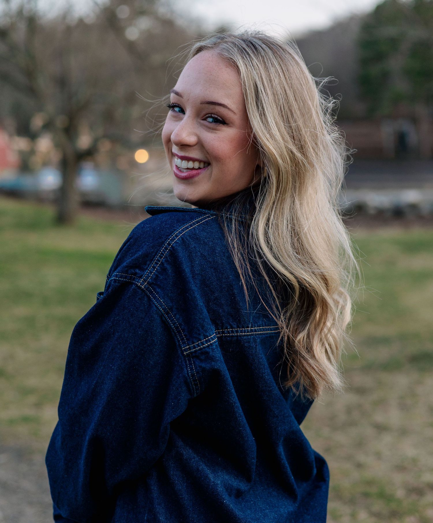 Smiling blonde woman in dark denim jacket looking over shoulder outdoors in park setting.