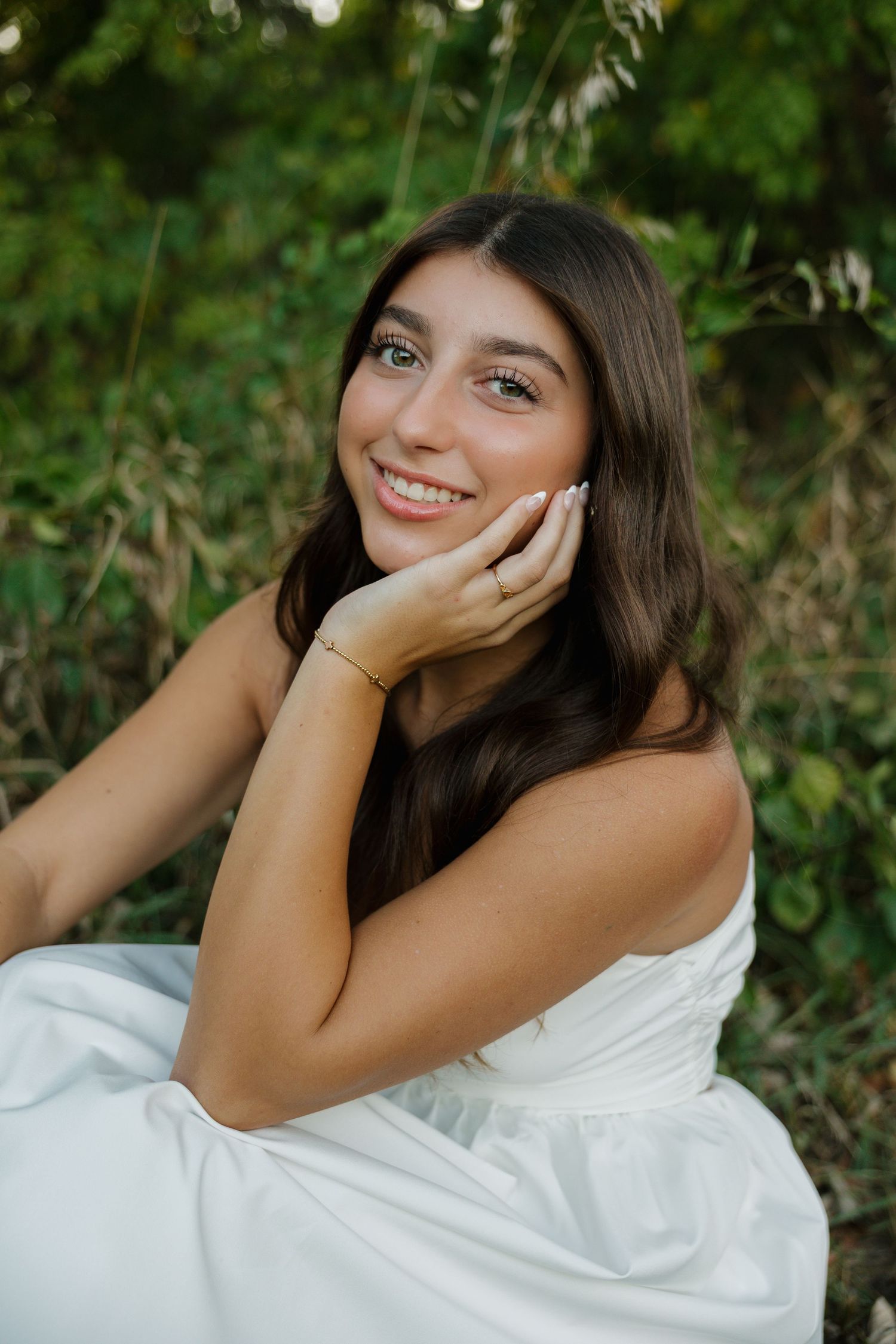 Young woman in white strapless dress smiling outdoors, resting chin on hand with green foliage background.