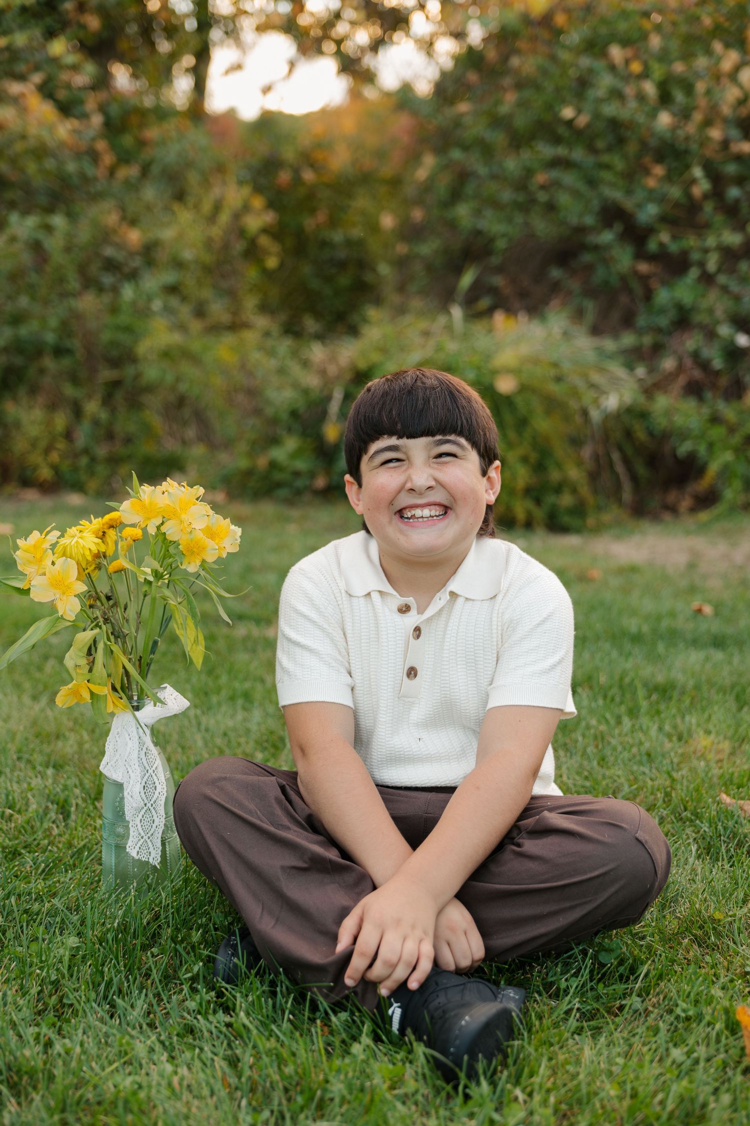 Smiling boy sitting cross-legged on grass with yellow flowers beside him in a lush green garden.