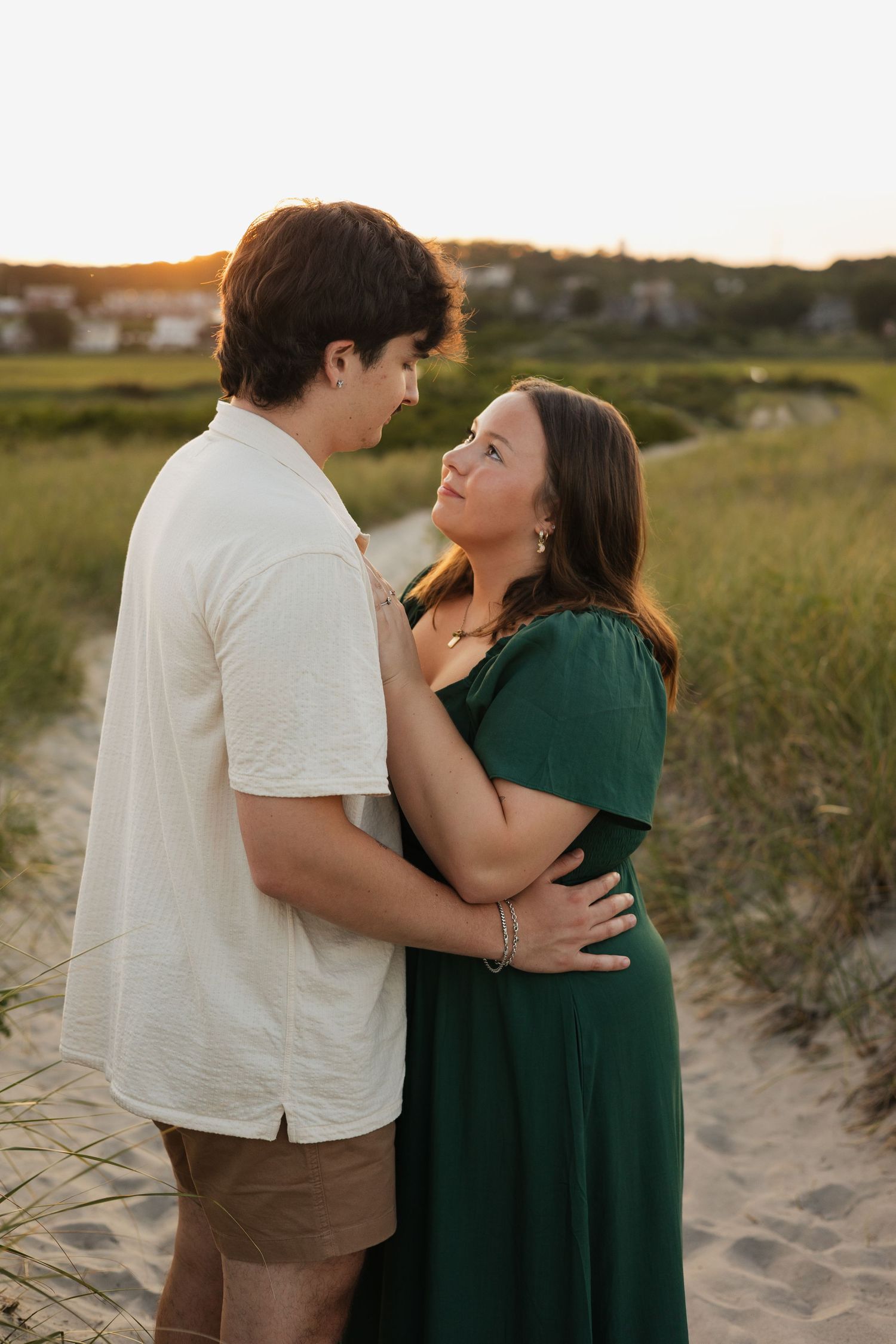 Couple embracing at golden hour on a sandy beach path, woman in green dress gazing up at man in white shirt.