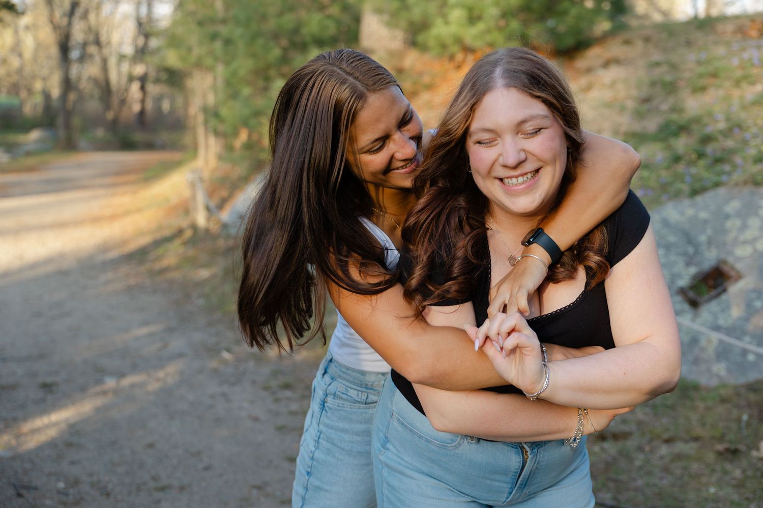 Two young women laughing and hugging outdoors in a sunny park setting.