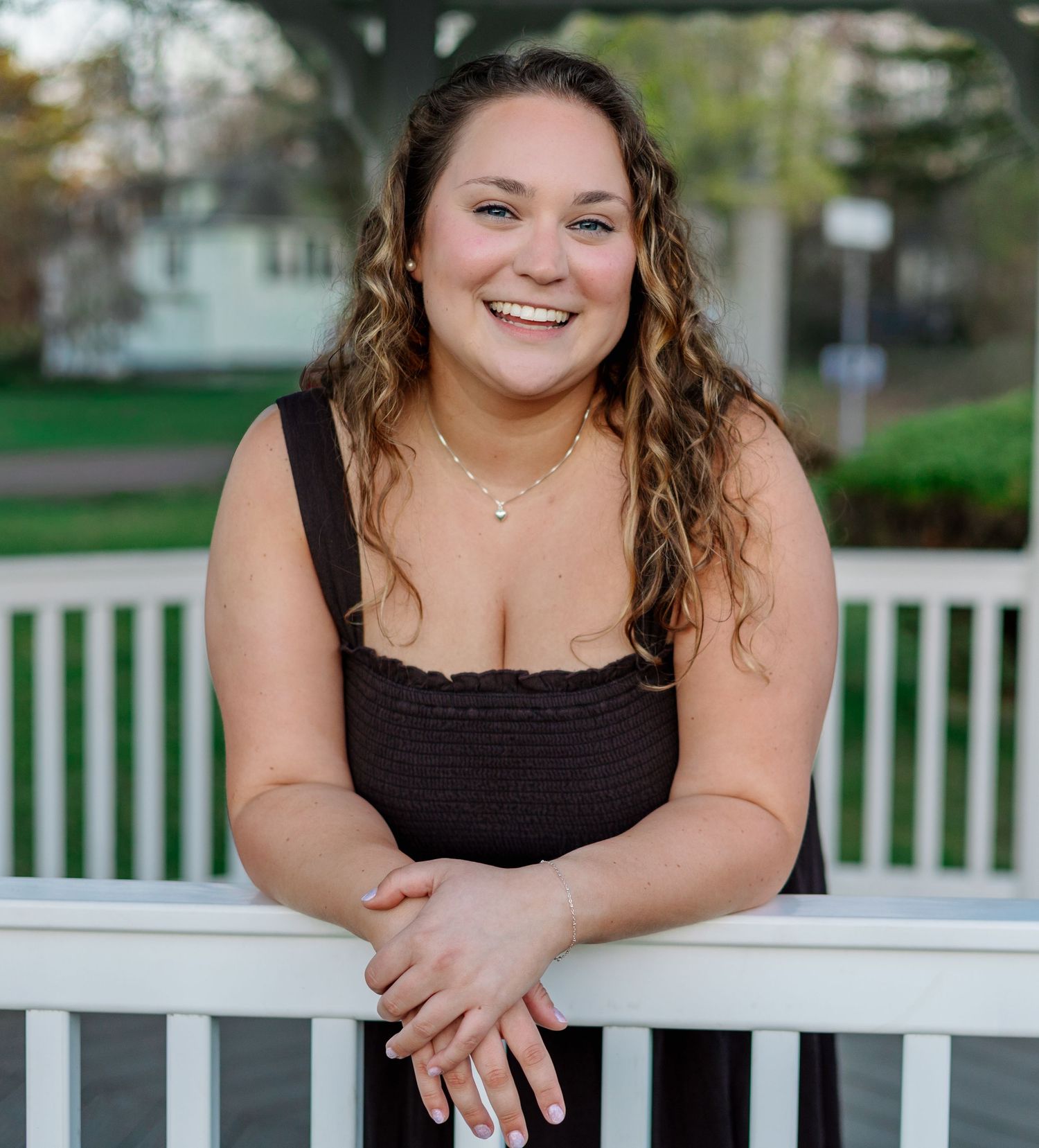 Smiling young woman in black sundress leaning on white railing outdoors during golden hour portrait session.