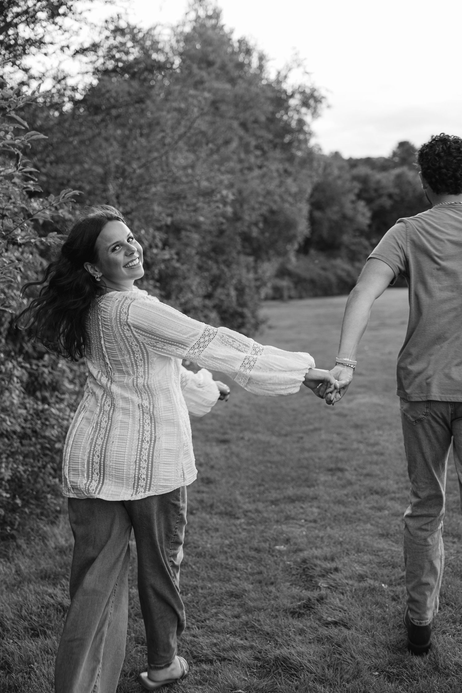 Black and white photo of a smiling woman holding hands with a man while walking through a lush green park.