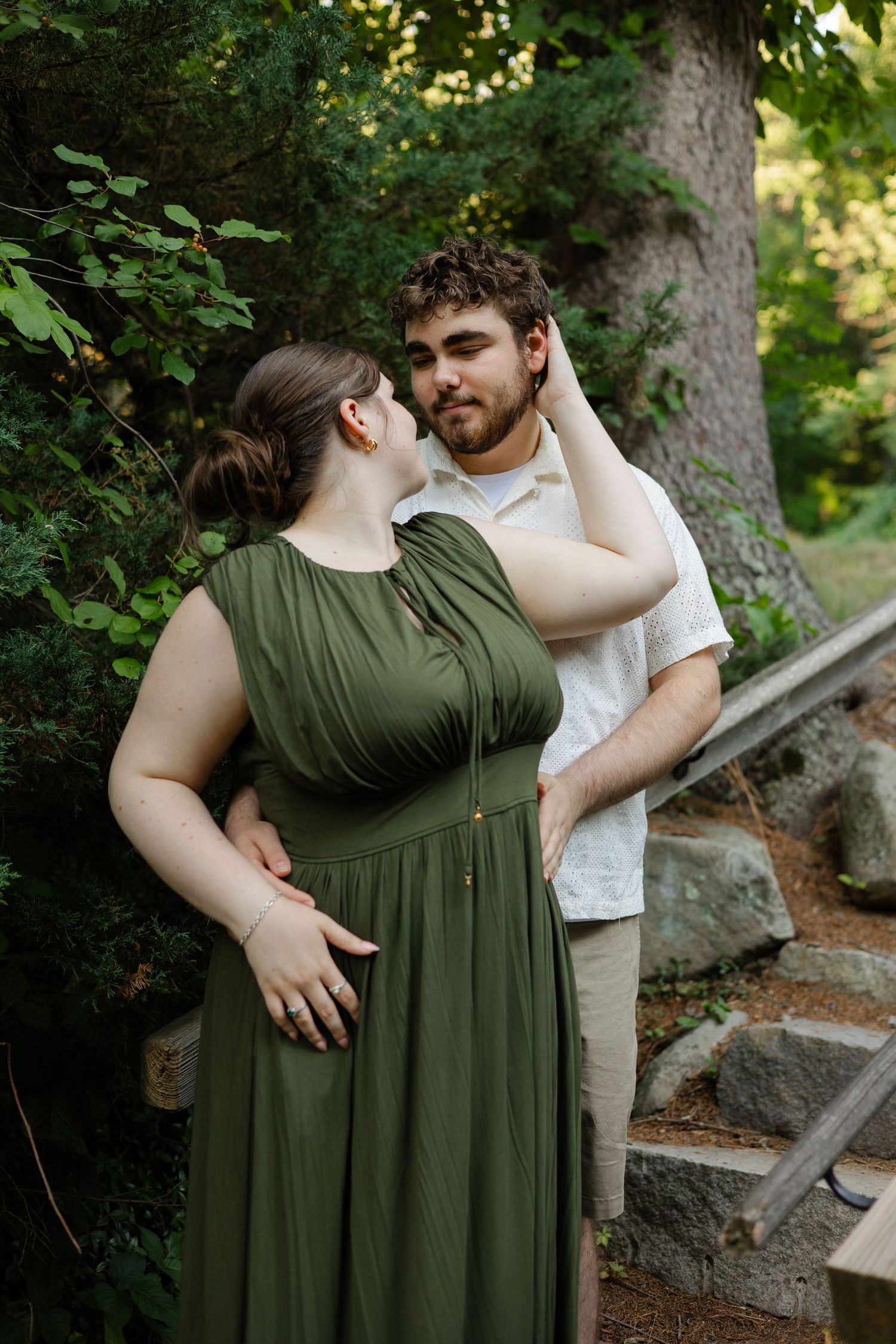 Couple in green dress and white shirt share a romantic moment outdoors near stone steps and lush trees.