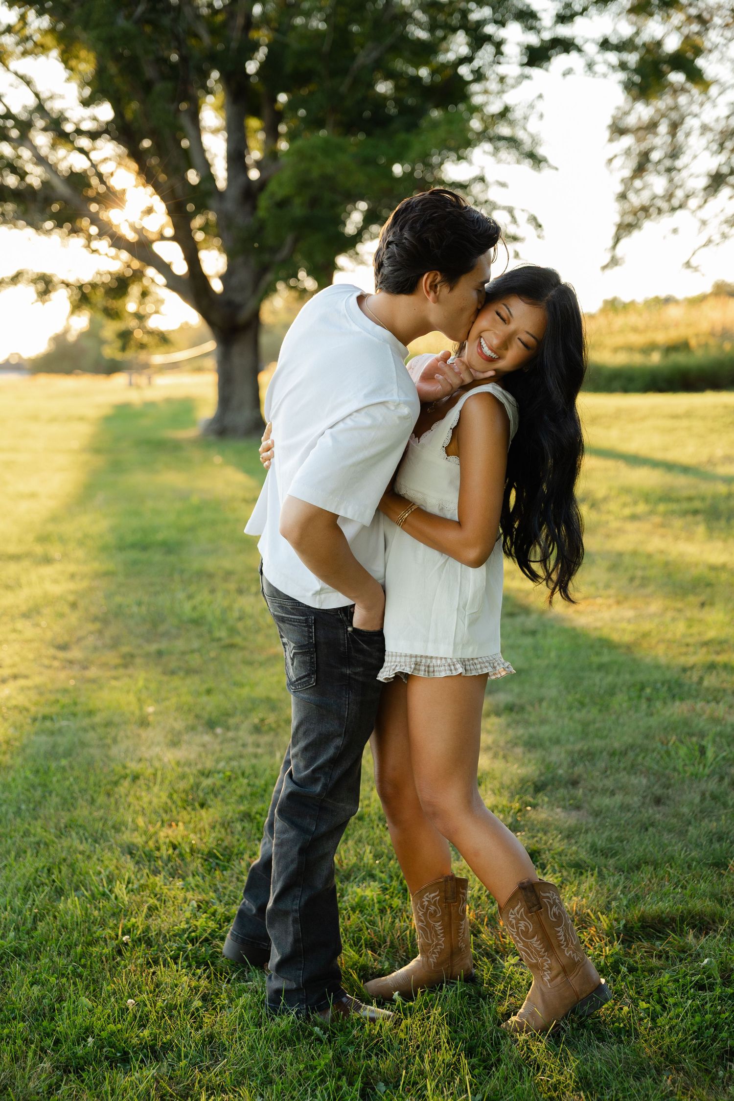 Couple embracing outdoors at golden hour, woman in white dress and cowboy boots smiling as man kisses her cheek.