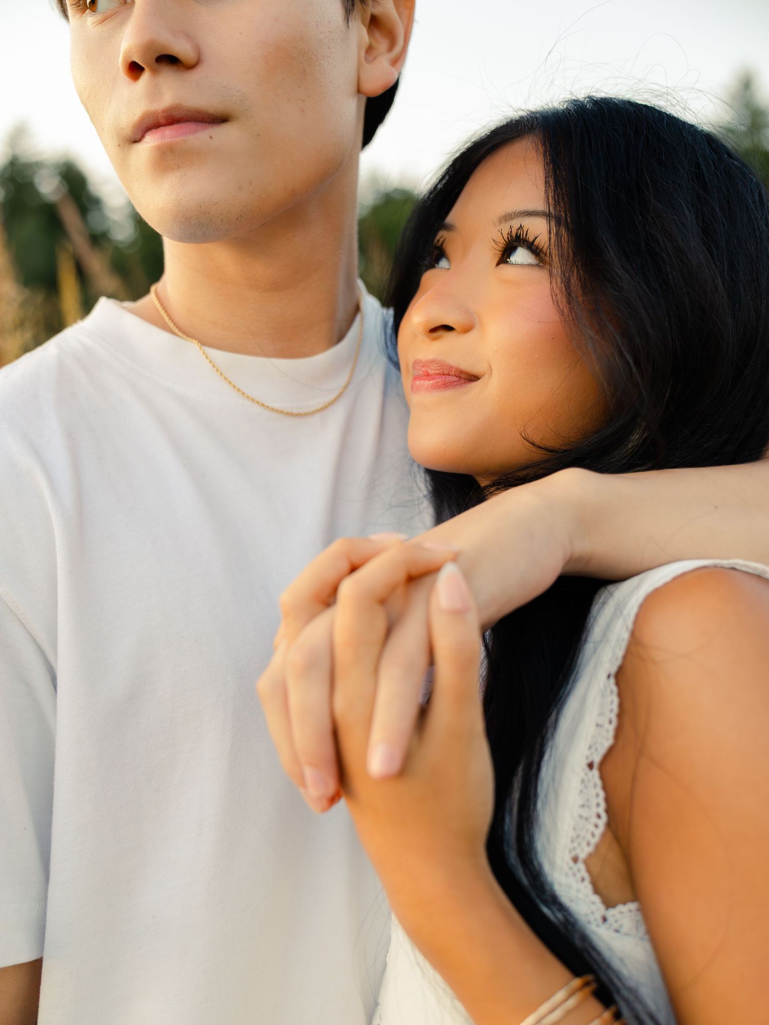 A young Asian couple embracing outdoors, woman in white dress gazing up lovingly at man in white t-shirt.