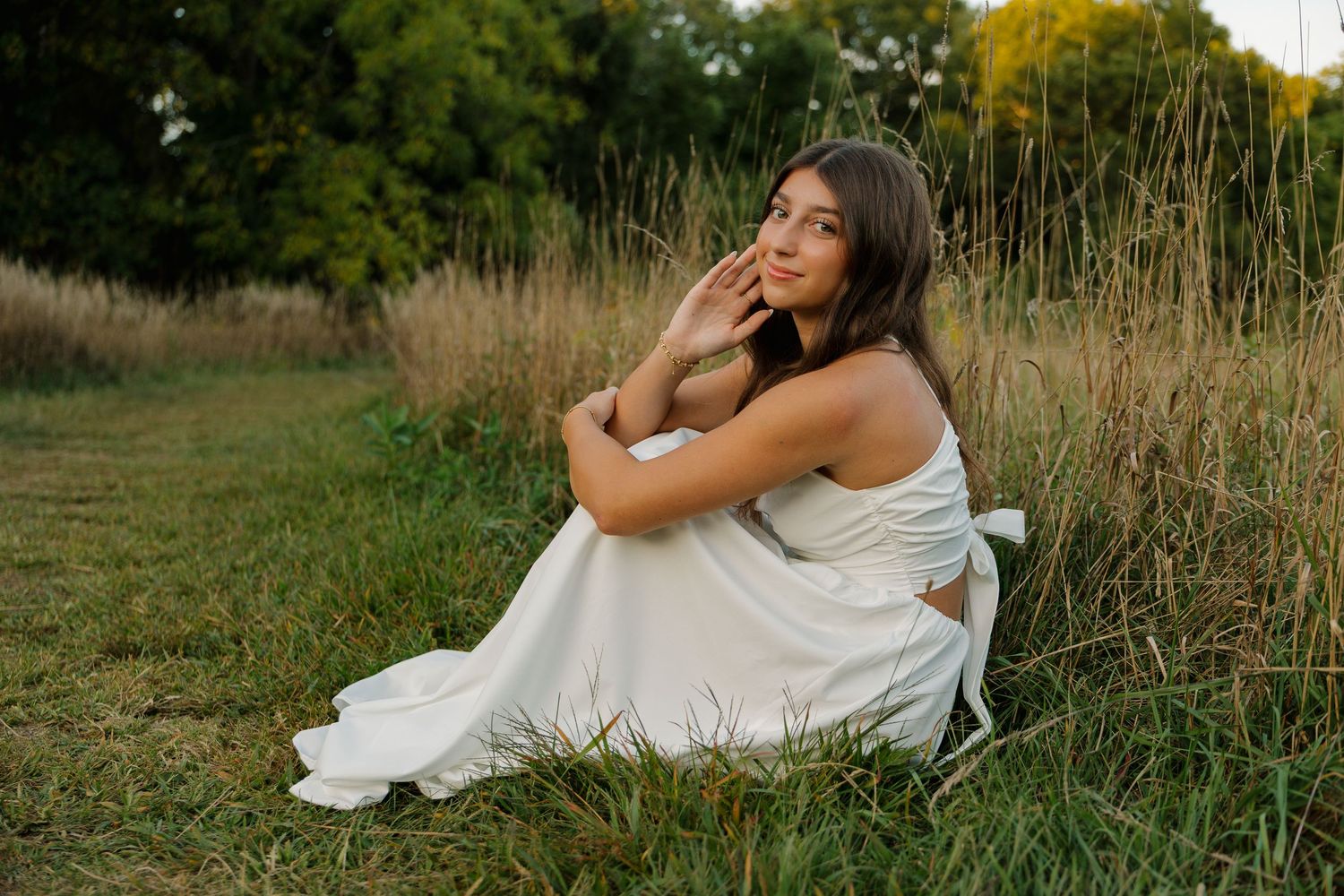 Young woman in white dress sitting gracefully in a grassy meadow with trees in background during golden hour.
