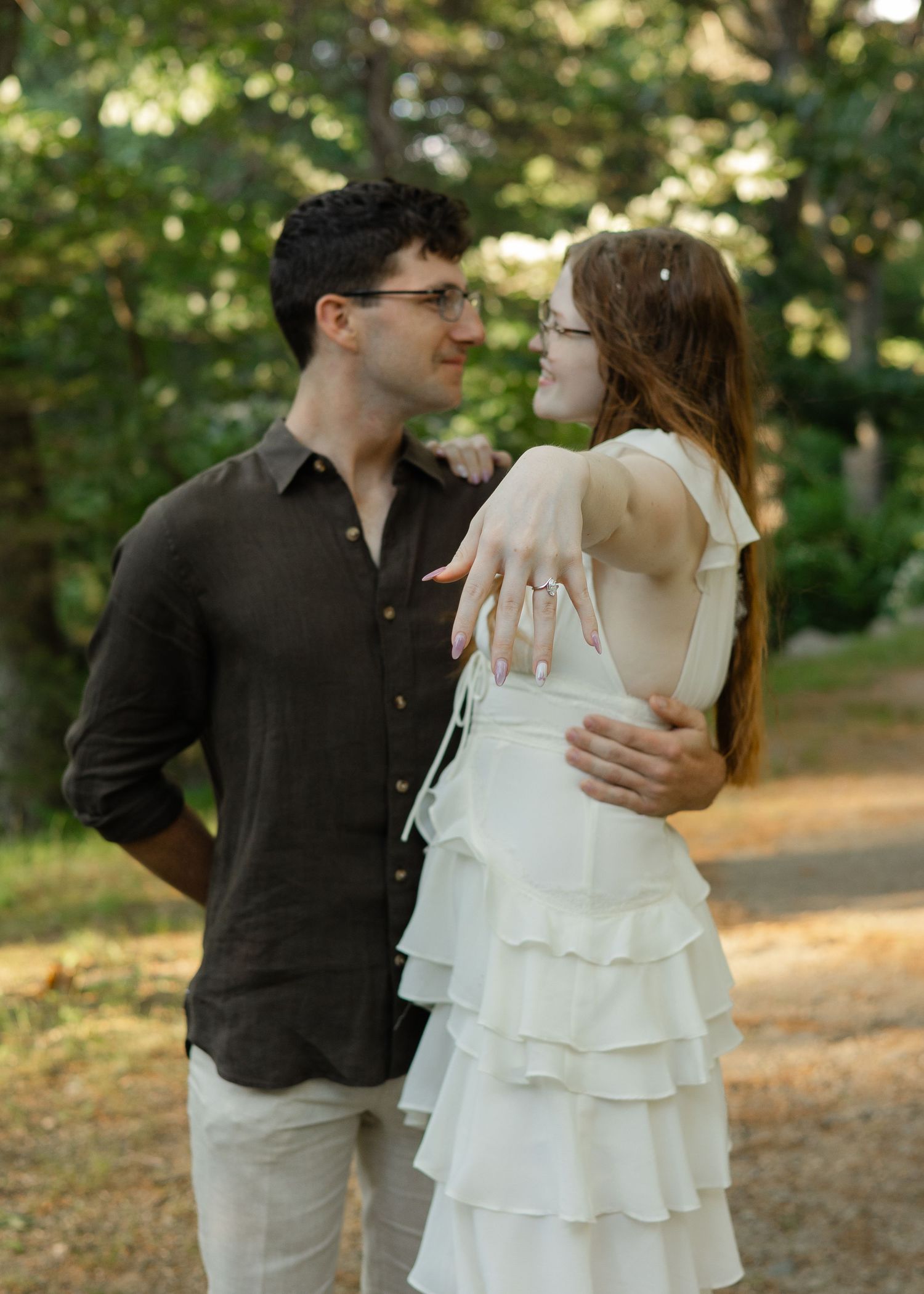 Couple in outdoor engagement photo, woman in white ruffled dress showing ring, man in dark shirt embracing her.