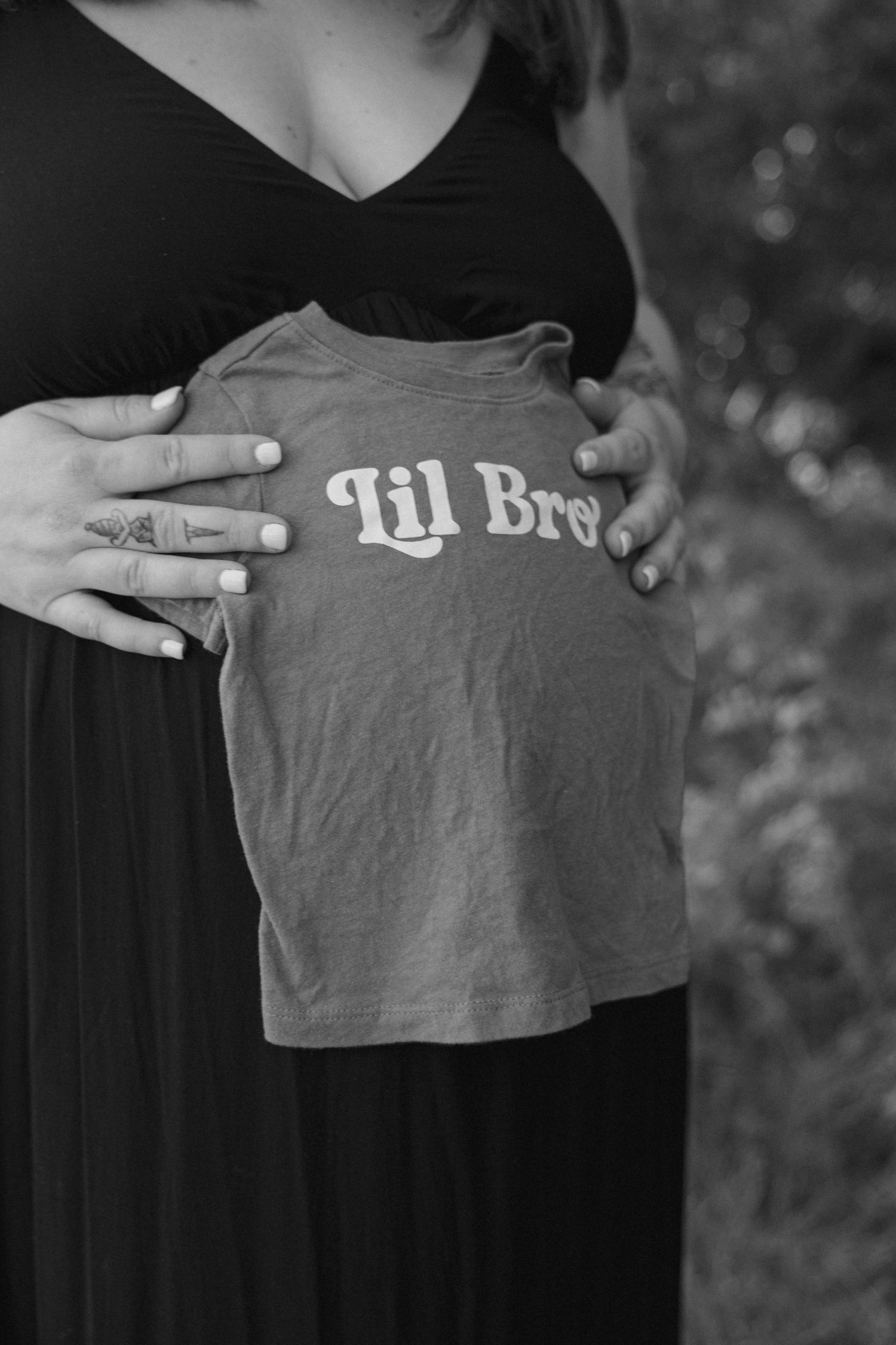 Pregnant woman holding a 'Lil Bro' shirt over her baby bump in a black and white maternity photo.