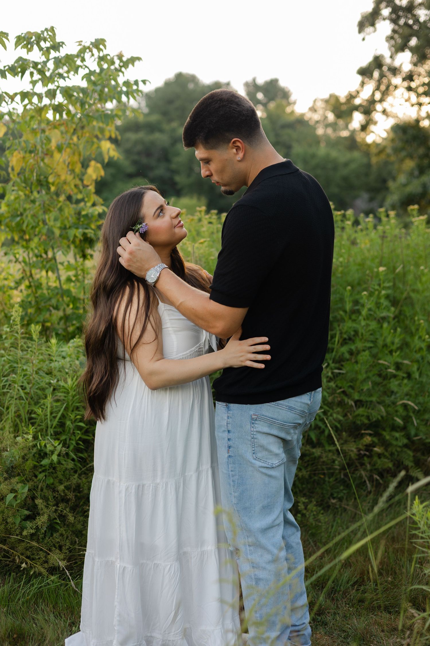 A couple embraces in a lush green field, woman in white dress gazes up at man in black shirt and jeans.