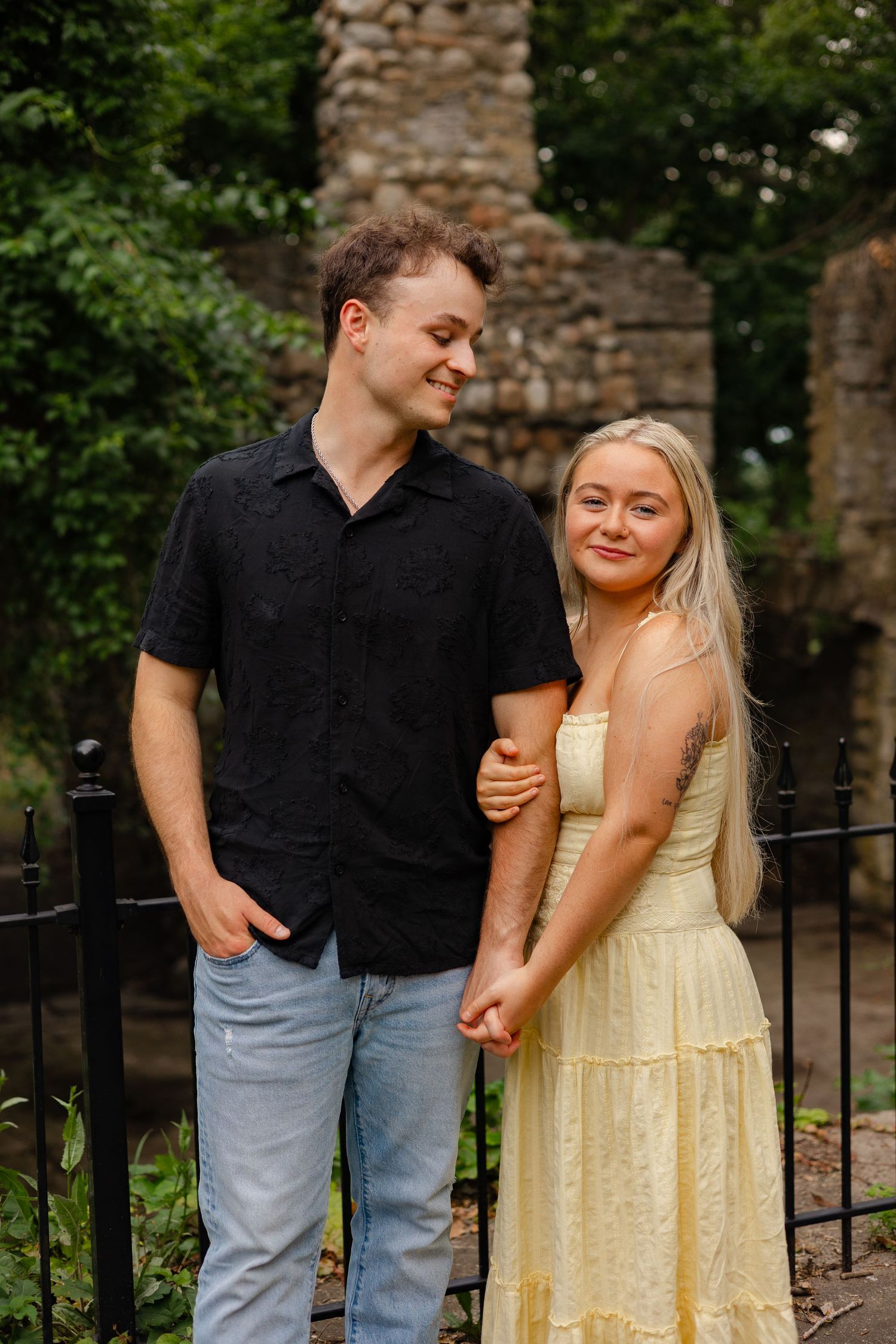 A young couple holds hands and smiles outdoors near a black iron fence with lush greenery behind them.