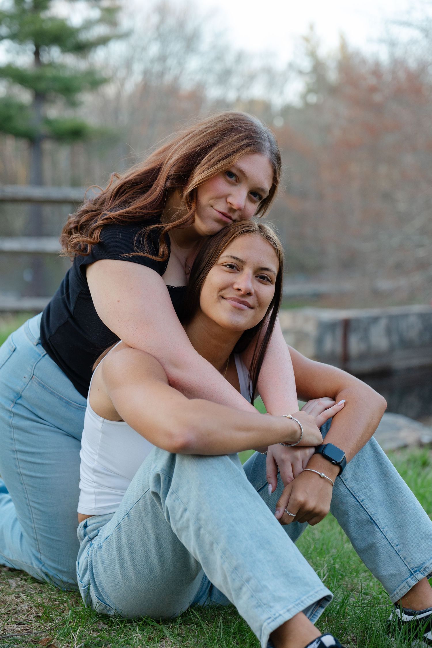 Two young women sitting on grass, one hugging the other from behind.