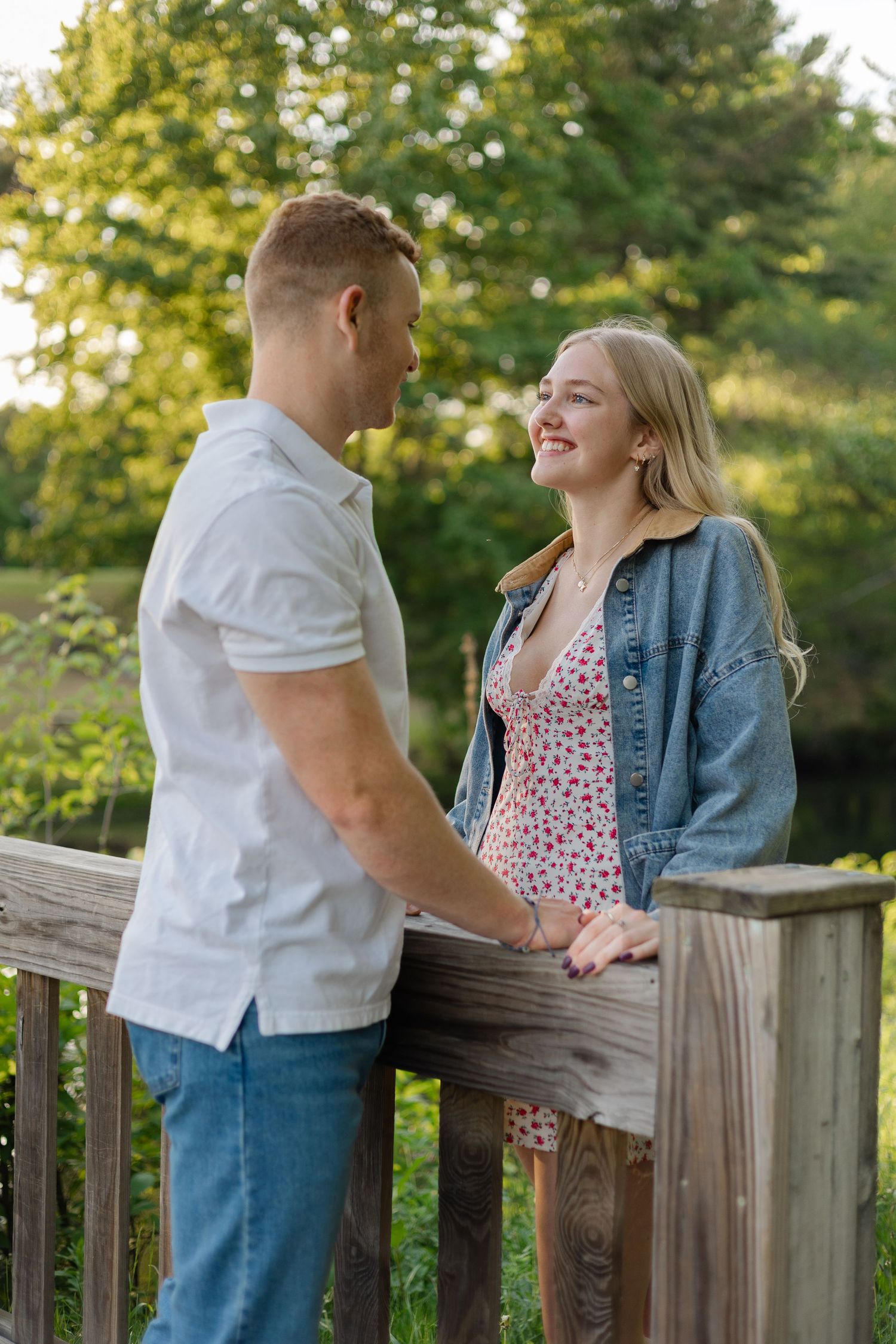 A smiling couple holds hands on a wooden bridge surrounded by lush green trees in warm golden hour light.
