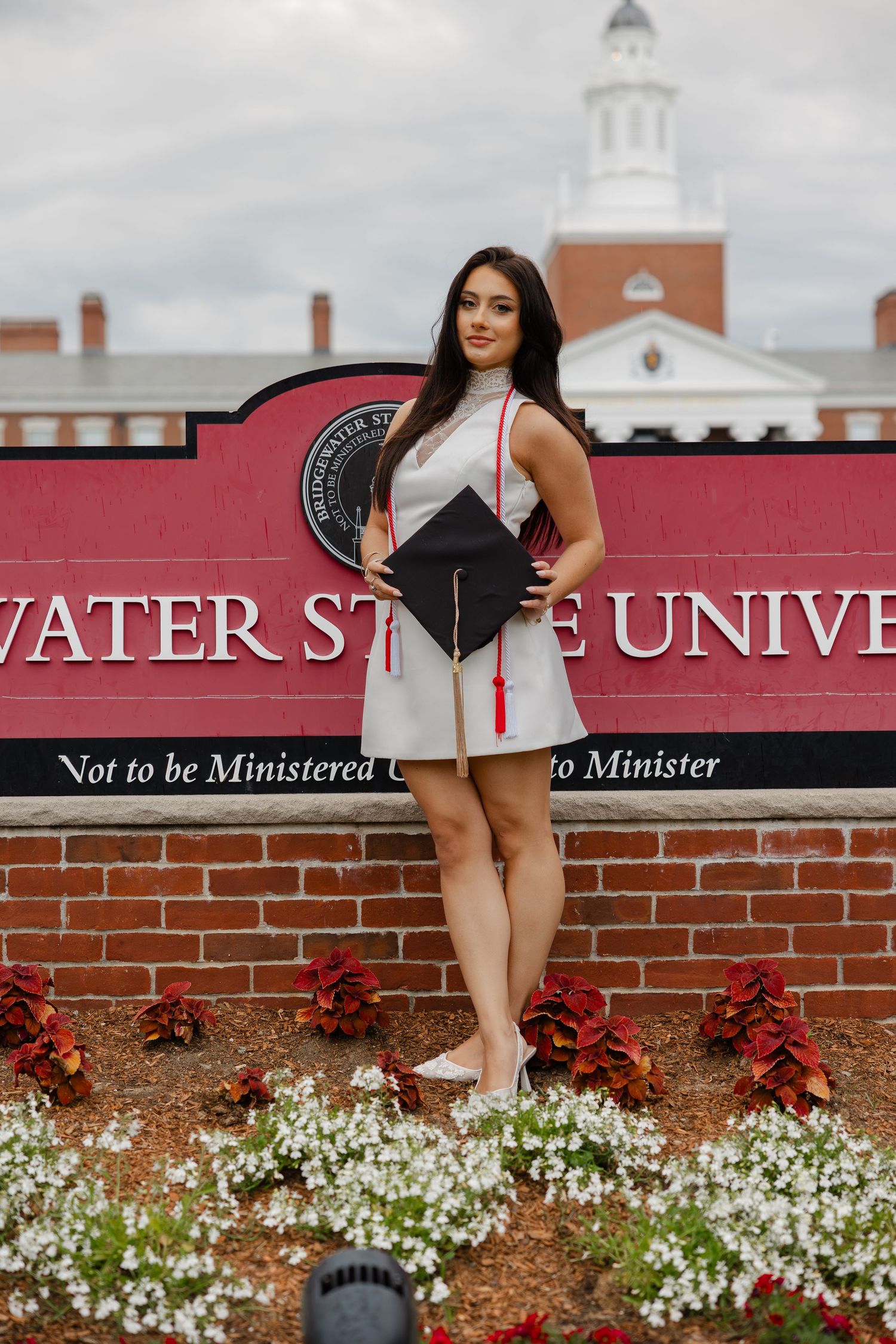 Female graduate in white dress holding cap in front of Clearwater State University entrance sign.