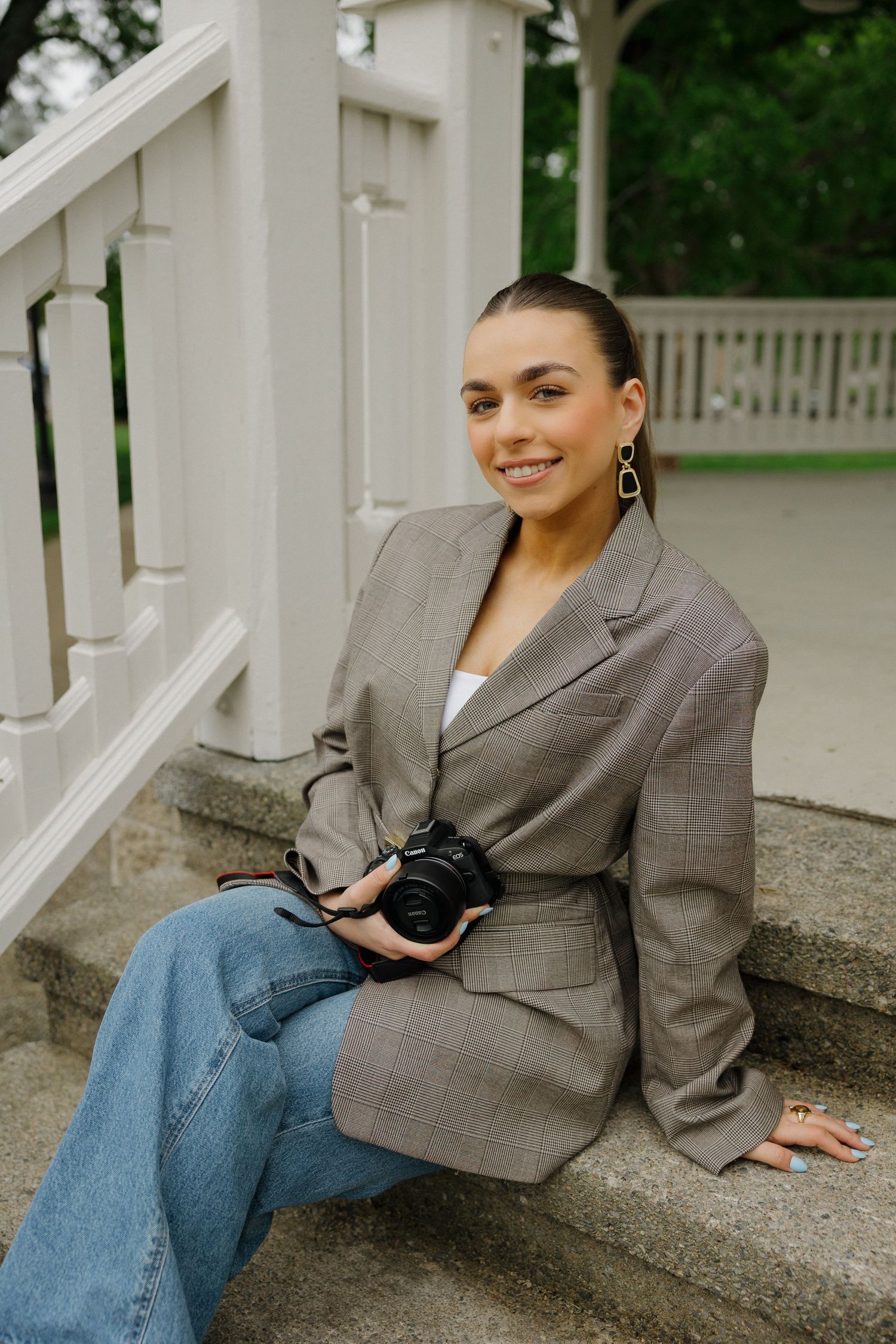 A smiling female photographer sits on stone steps holding a Canon camera in a gray belted blazer and jeans.
