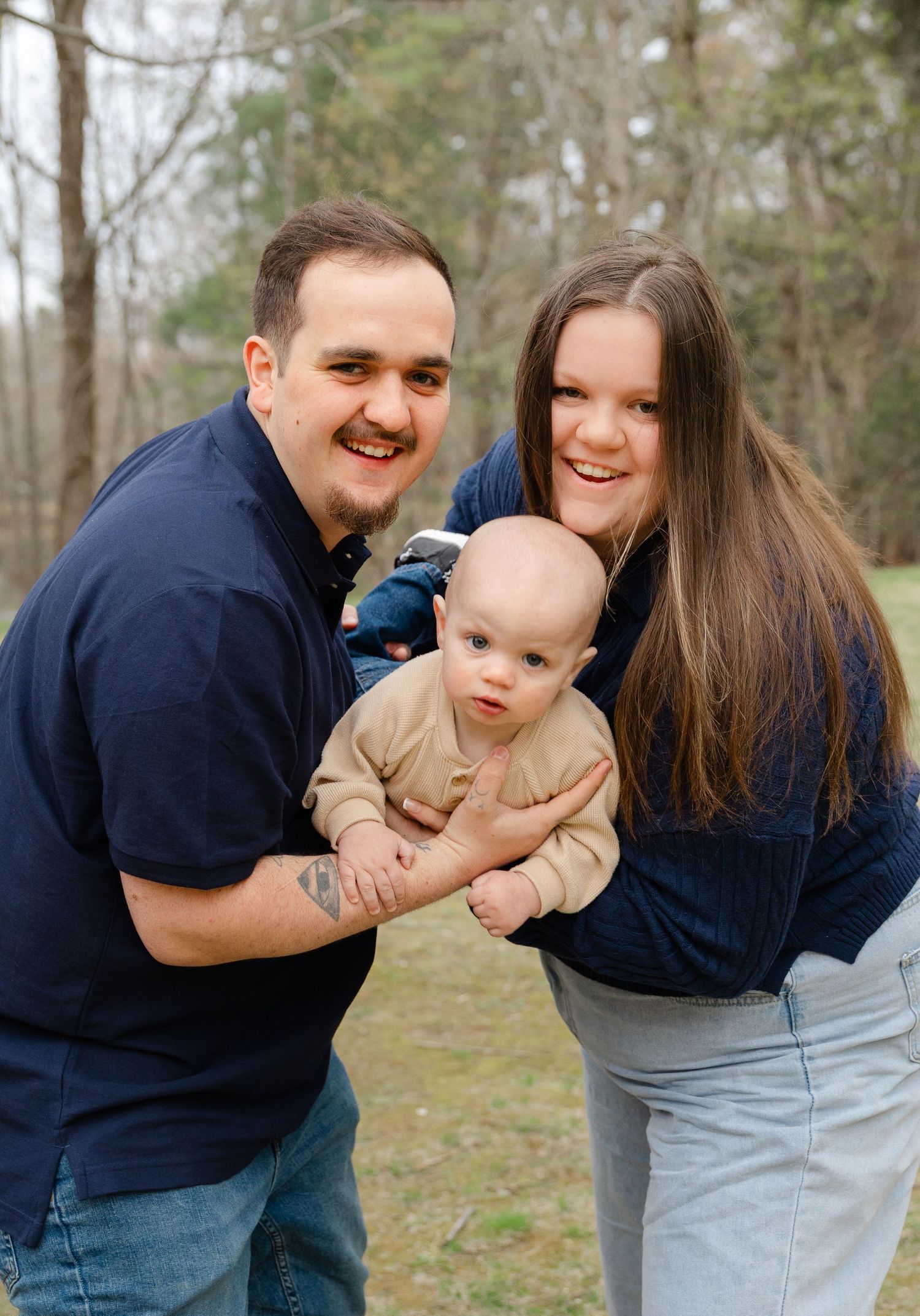 Happy young family with a baby outdoors, parents in navy shirts smiling, trees in background.