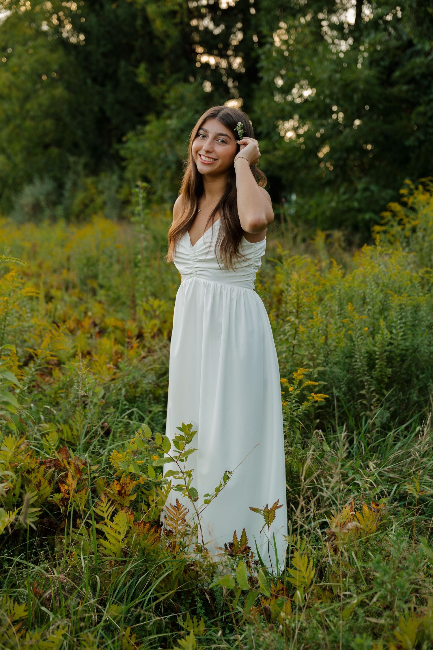 Young woman in white maxi dress standing in wildflower field, smiling outdoors at golden hour.
