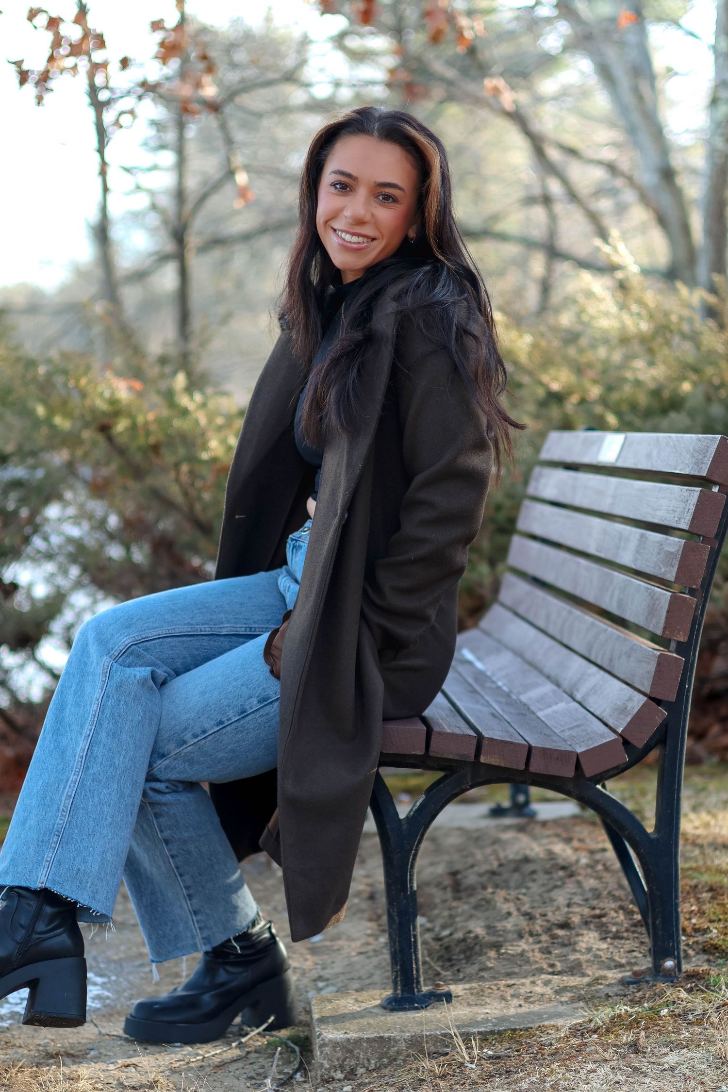 Smiling young woman in dark coat and flared jeans sitting on a park bench surrounded by autumn foliage.