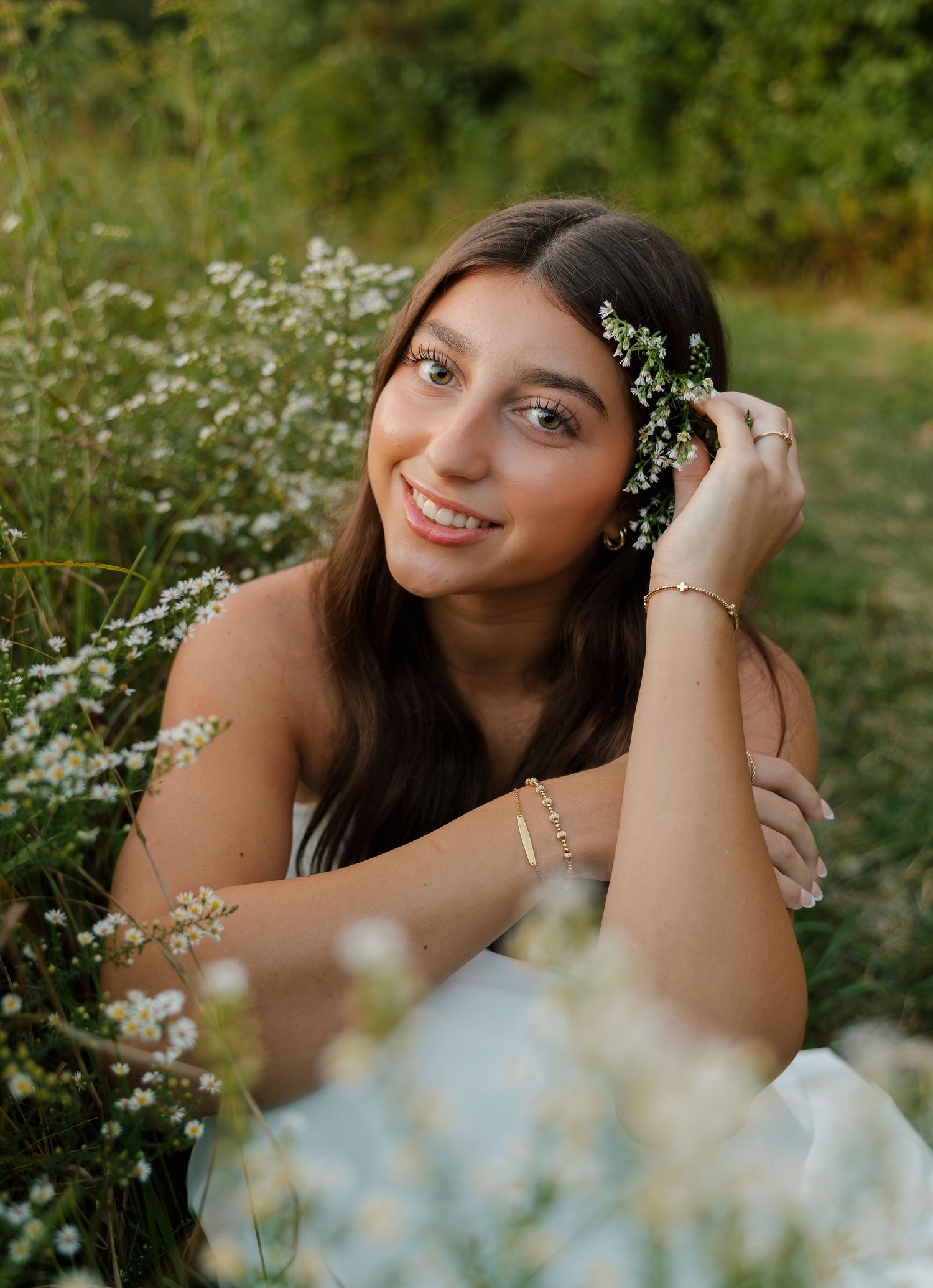 Young woman smiling in a wildflower field, holding white flowers in her hair, wearing a white dress and gold jewelry.