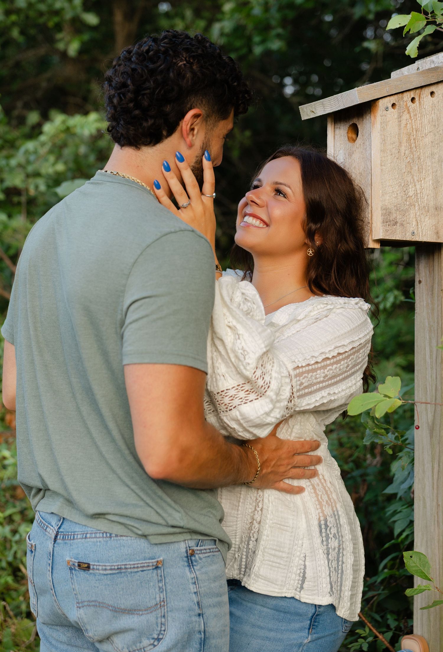 A smiling woman in a white lace blouse embraces a man in a green shirt outdoors near a wooden birdhouse.