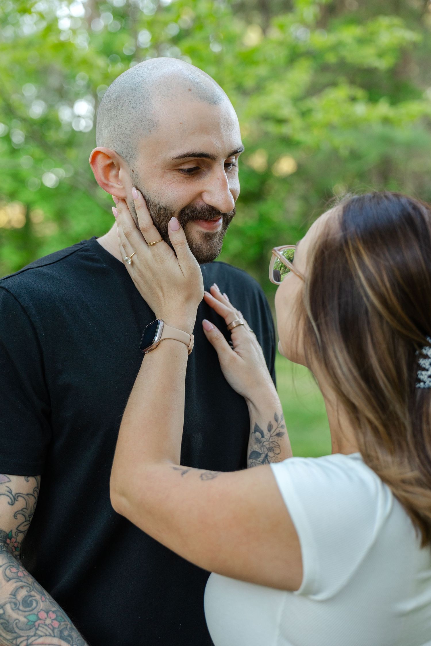 Couple sharing a tender moment outdoors, woman touching mans bearded face, both smiling, green trees background.