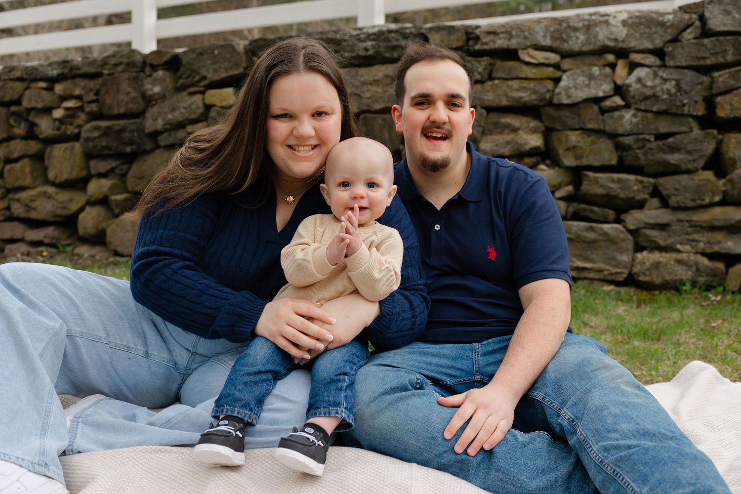 Happy young family with baby sitting together on white blanket against rustic stone wall outdoors.